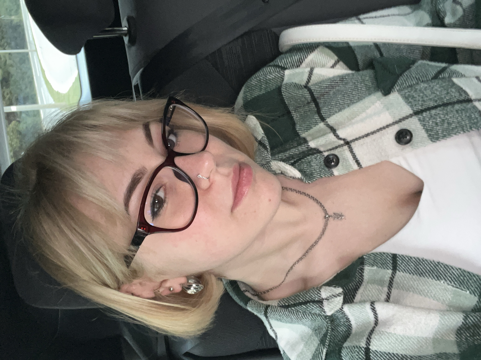 The image is a close-up, selfie-style photograph of an individual, rotated approximately 90 degrees counter-clockwise from an upright orientation. The subject, appearing to be female, has blonde hair styled with bangs and is wearing dark-rimmed glasses, possibly with a subtle red hue on the frames. She looks directly at the camera with a neutral expression.
Notable facial features include a small silver hoop piercing on her left nostril and two visible piercings on her right ear: a small stud and a larger, ribbed silver earring. She is wearing a green, white, and dark grey plaid flannel shirt over a plain white t-shirt. Around her neck, a thin silver chain necklace is visible, featuring a small, stylized pendant that resembles an angel or winged figure.
The setting appears to be the interior of a vehicle, likely a car, indicated by the dark grey or black fabric of a headrest and seat visible behind and to the side of the person. Through a window in the upper background, a blurred outdoor scene can be discerned, showing green foliage and a soft-focus white or light-colored architectural element. The external background is generic and offers no specific visual cues to identify the location as Rafailovići, Montenegro. The lighting suggests it is daytime. No visible text is present in the image.