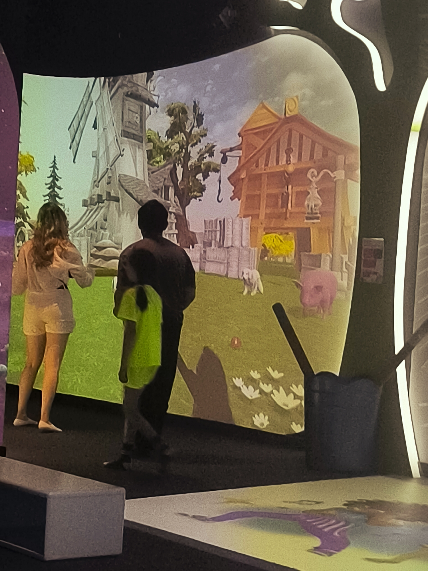 Two individuals are standing in front of a projected image of a rural scene. The projection displays a windmill on the left and a wooden building with a yellow haystack on the right, set against a cloudy sky. In the foreground of the projection, a pink pig and a white dog are visible. The individuals are positioned on a dark floor, with one person, a woman wearing a white outfit, facing the projection. The other person, a man wearing a dark shirt and a neon green t-shirt draped over his shoulder, is also facing the projection, with his back to the camera. A dark cylindrical object is located to the right of the man. The floor also has another projection of a purple figure. There is no indication of the Qarağandı city administration or Kazakhstan in the image.