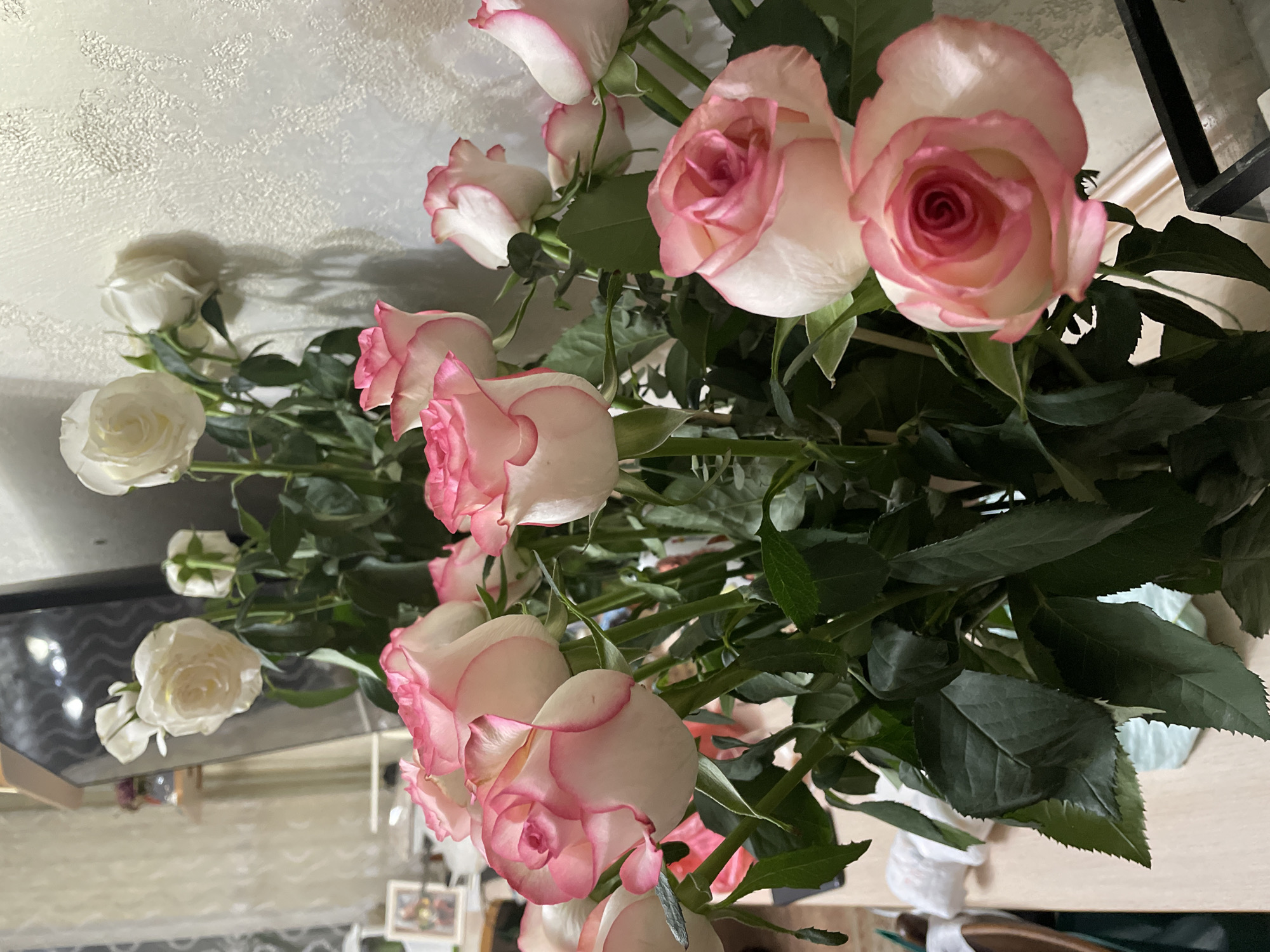 A bouquet of roses is displayed in a room. The roses are predominantly white with pink edges, some are fully bloomed, while others are still buds. The bouquet is arranged with lush green leaves. The background features a textured, light-colored wall and a reflective surface, possibly a table or mirror, showing a blurred reflection of the room, including what appear to be framed pictures and some white tissue paper. The lighting suggests an indoor setting, possibly during the daytime. The overall impression is one of a decorative arrangement, likely intended for a special occasion or as a gift. There is no visible text in the image.