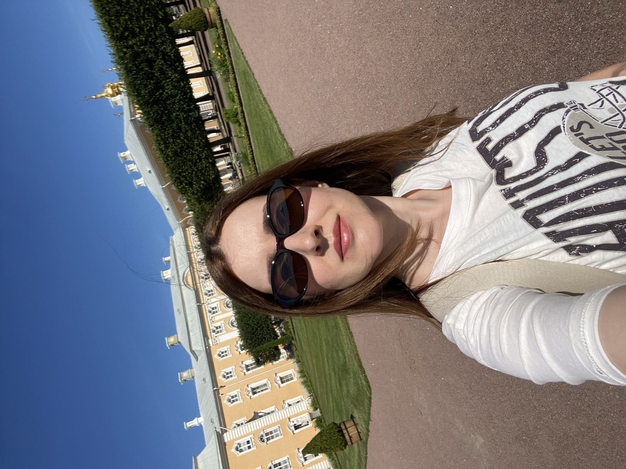 A woman with long brown hair and sunglasses is taking a selfie in front of a yellow building with a grand facade. She is wearing a white t-shirt with a graphic design. The background features a manicured lawn, trimmed trees, and a clear blue sky. The architecture suggests the location is the Peterhof Palace in Peterhof, Russia, a suburb of Saint Petersburg. The day is bright and sunny, indicating it is likely daytime.