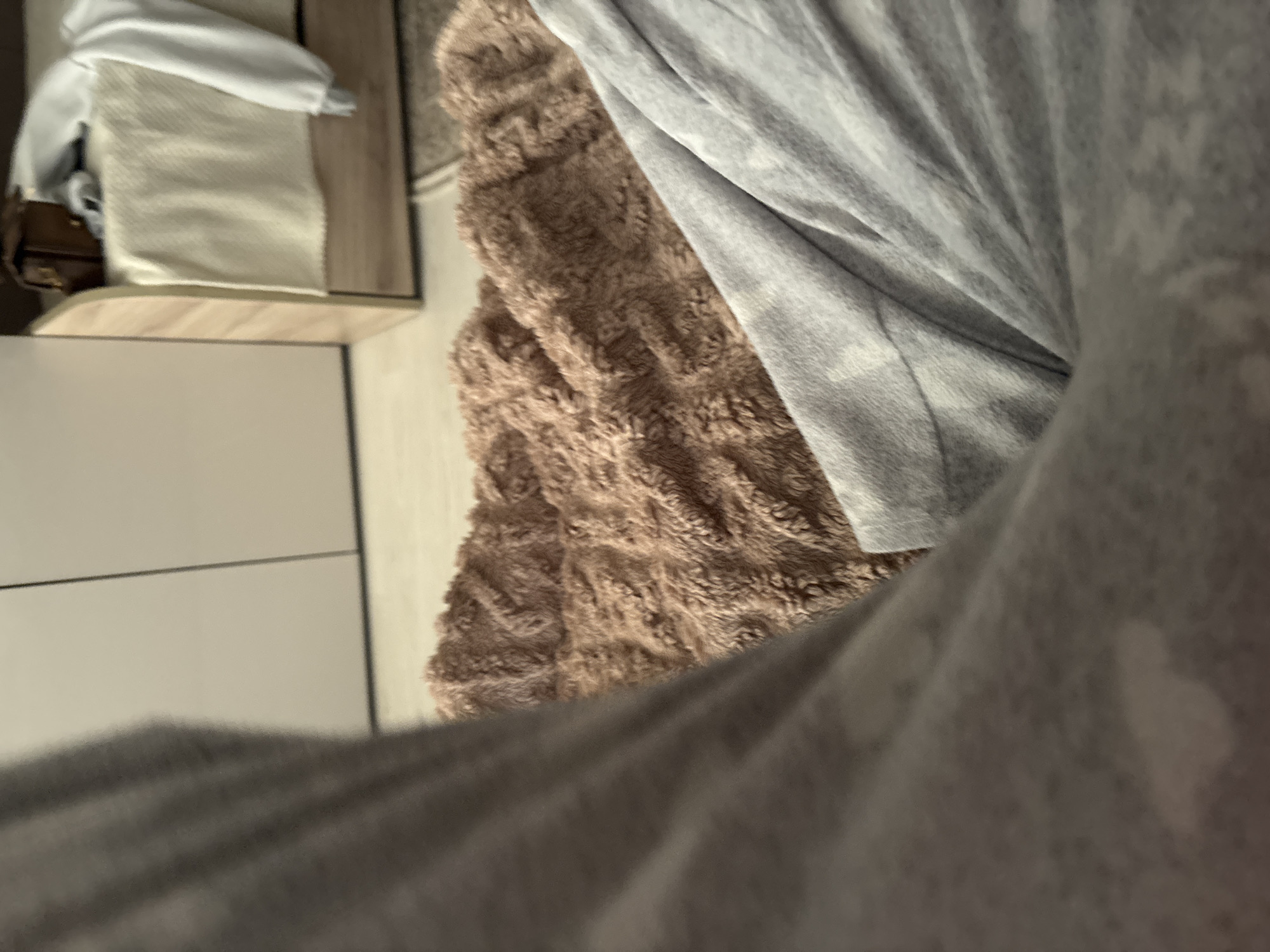 The image shows a close-up, low-angle view of a person's legs covered by soft, gray loungewear. In the background, a textured, brown blanket is visible, and further back, a bed with white bedding and a small brown handbag. The environment appears to be a bedroom. The specific location within the city of Unknown, Russia is not discernible from the provided image.