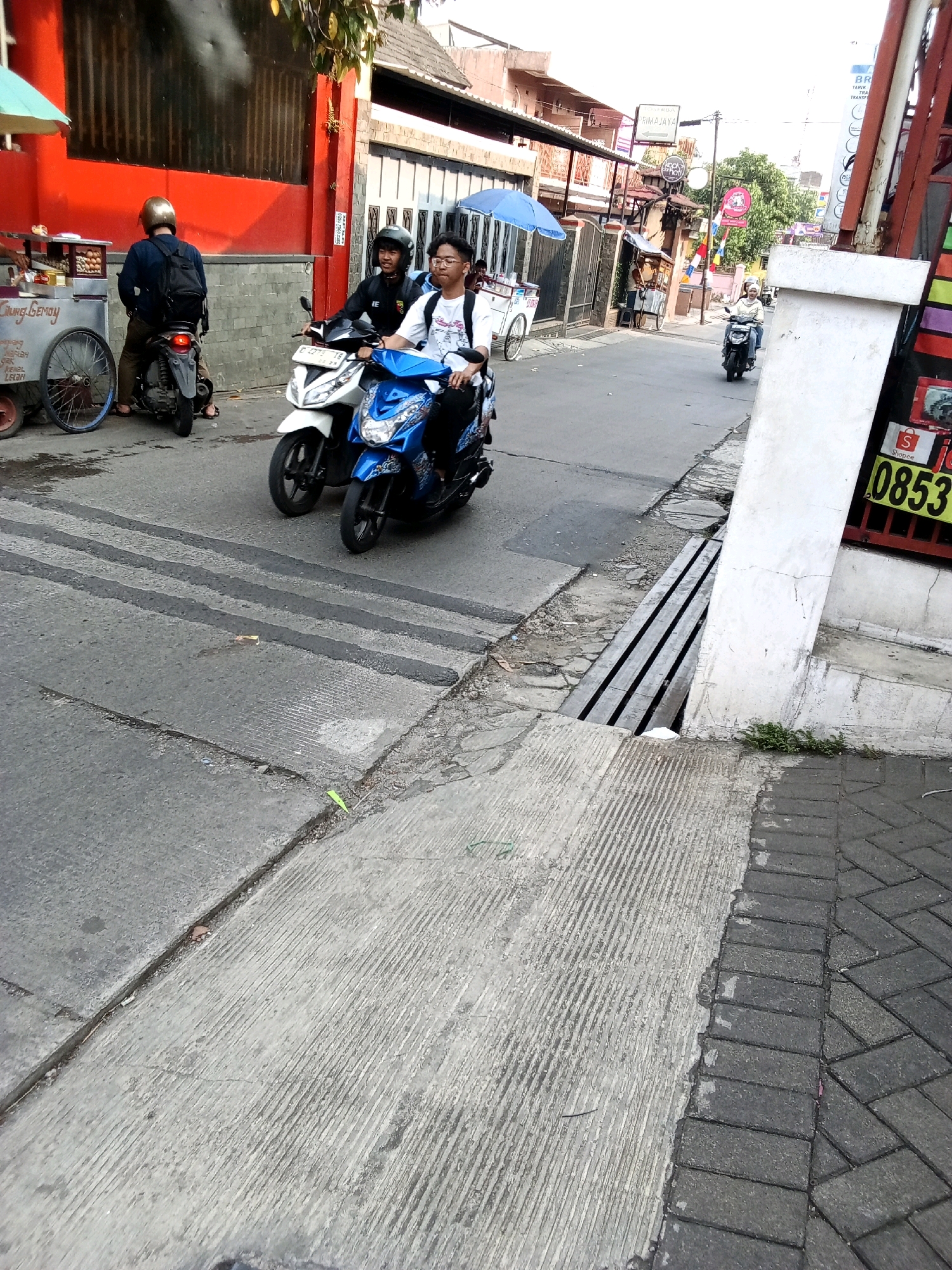 The image depicts a street scene in Bandung City, Indonesia, with a focus on a road and sidewalk area. Two young men on a blue scooter are in the foreground, moving away from the camera. The driver is wearing glasses and a white t-shirt, while the passenger behind him wears a black jacket and a helmet. To their left, another scooter with a rider in a helmet and a backpack is parked.
The road surface is asphalt, with prominent speed bumps visible. A concrete sidewalk runs along the right side of the road, bordered by brick pavers. A drainage grate is situated between the road and the sidewalk. In the background, buildings line the street, some with shops and stalls. One stall, to the left of the frame, appears to be selling food. The overall impression is a bustling urban environment, typical of a city in Indonesia.