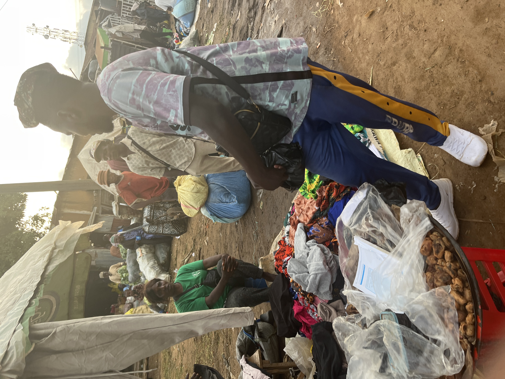 The image depicts an outdoor market scene in Gura Top, Nigeria. In the foreground, a man wearing a patterned t-shirt and blue athletic pants with a yellow stripe is facing away from the camera, looking to his right. He has a black bag slung across his chest. To his left, a woman in a green t-shirt is sitting, looking towards the camera. In front of her is a pile of colorful fabrics, some of which are wrapped in plastic. A red stool supports a tray containing what appears to be food items covered in plastic. Further back, other people are visible, some tending to goods, including produce, and others browsing. Large bags, possibly filled with merchandise, are stacked in the background. The environment is dusty and appears to be a bustling commercial area, with various structures and a telecommunications tower visible in the distance.