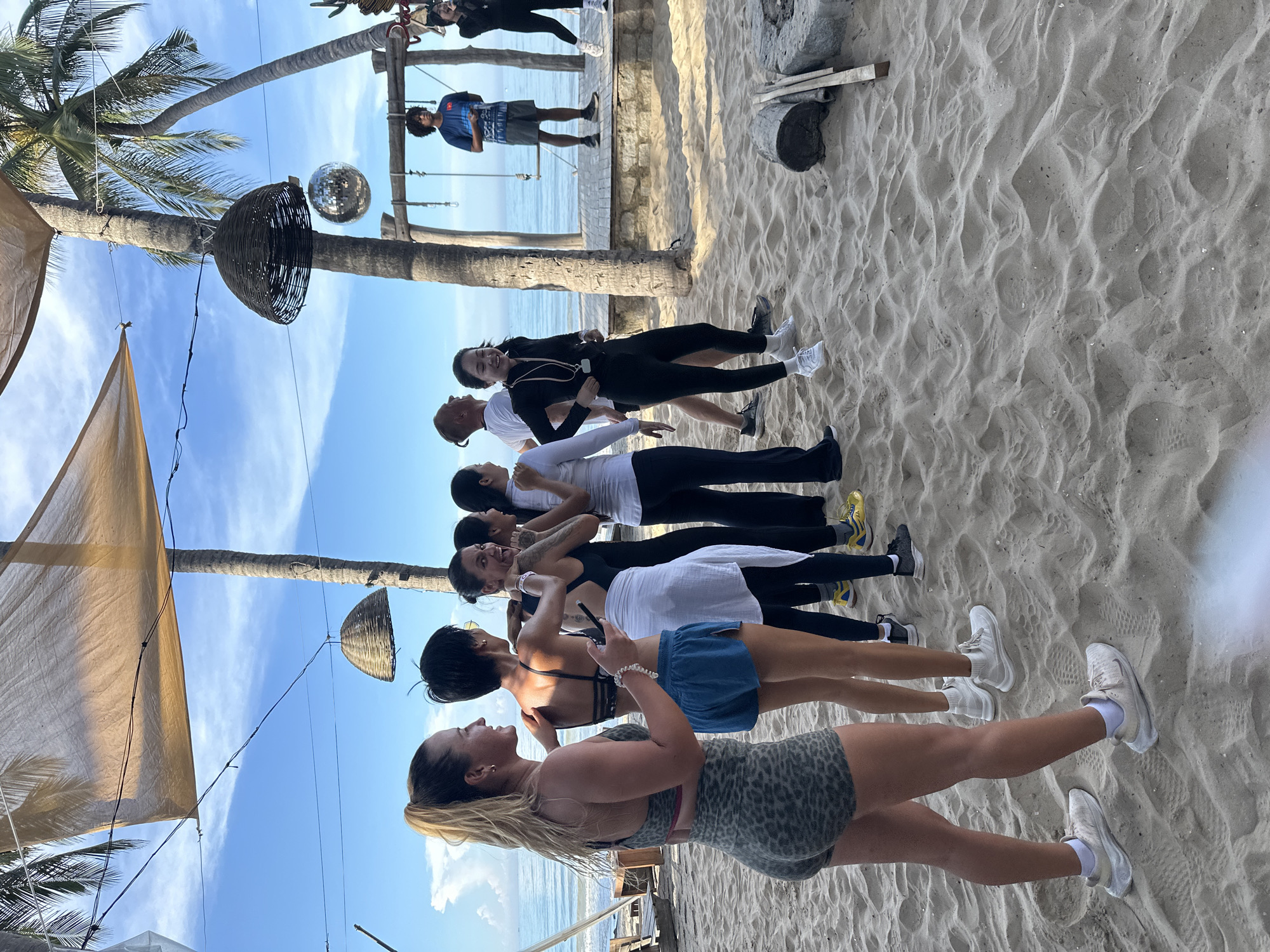 A group of nine individuals, appearing to be young adults, are gathered on a sandy beach, likely in Mũi Né, Vietnam. The scene is outdoors, with palm trees and the ocean visible in the background. The sky is blue with some clouds, and the sun appears to be shining.
In the foreground, a woman with blonde hair tied in a ponytail is facing to the left, her body angled towards a group of other individuals. She is wearing leopard-print athletic shorts and a matching sports bra. To her right, a person with short black hair is wearing a black sports bra and blue athletic shorts. They are holding a dark object, possibly a phone, in their right hand and looking towards the left.
The group continues to the right with several individuals in athletic wear, some in black leggings and tops, and others in white shirts and dark pants. They are all facing generally in the same direction, suggesting they are participating in an activity together, possibly a fitness class or a dance session, given the presence of a disco ball hanging from a palm tree.
Further in the background, two men and a woman are standing on a raised wooden platform. One man wears a blue t-shirt and shorts, while the other wears a white t-shirt and dark pants. The woman on the platform is wearing a dark outfit and white sneakers. There is a swing set structure behind them.
The beach is sandy, with footprints visible. Some logs and stones are present on the sand. Overhead, a light-colored canopy is stretched between poles, providing shade. The overall impression is a casual, active gathering on a tropical beach.