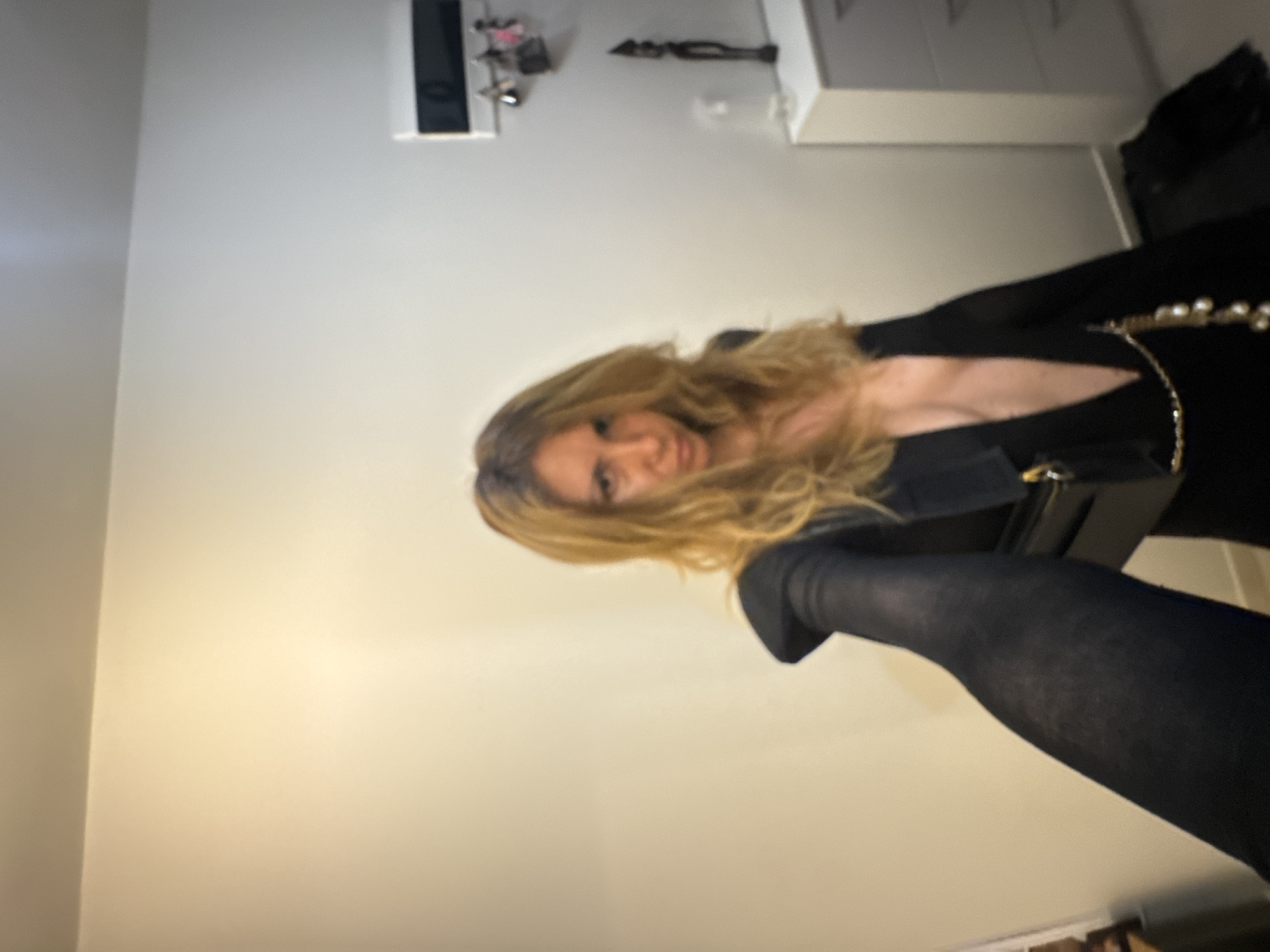 A woman with blonde, wavy hair and a plunging neckline is taking a selfie in what appears to be a dimly lit indoor environment. She is wearing a black garment that resembles a blazer or dress with wide sleeves and is holding a black purse with a gold chain and pearl embellishments. Her left arm, clad in black hosiery, is extended towards the camera. In the background, a wall with a light switch and key hooks is visible, along with a white dresser and a dark decorative object. The scene is situated within an apartment in Sofia, Bulgaria.