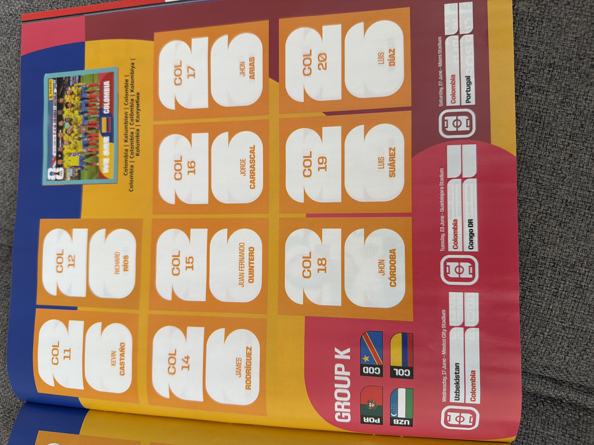 This is a photograph of a page from a sticker album, likely focused on a sports team or event, specifically showing stickers and information related to "COLOMBIA". The page is predominantly orange and yellow, with a blue header.
In the upper portion of the page, there is a small framed image of a soccer team wearing yellow jerseys. Text below this image reads "WE ARE COLOMBIA". Various stickers with the designation "COL" followed by a number (11 through 20) are arranged in rows. Each of these stickers has a number and a name printed on it. Some of the names visible include KEVIN CASTAÑO, RICHARD RIOS, JAMES RODRÍGUEZ, JUAN FERNANDO QUINTERO, JORGE CARRASCAL, JHON ARIAS, JHON CÓRDOBA, LUIS SUÁREZ, and LUIS DÍAZ.
Below the stickers, a section labeled "GROUP K" is visible. Within this section, there are representations of national flags for Portugal (POR), Congo (COD), and Uzbekistan (UZB), as well as the flag of Colombia. Below these flags are printed match schedules, including dates, locations, and participating teams such as Uzbekistan, Colombia, and Congo DR, and Portugal.
The image is taken from a slightly elevated angle, with the page lying flat on a textured, dark gray surface, possibly fabric such as upholstery or a carpet. The lighting appears to be from above, casting minimal shadows. There is no discernible information about the city of Zlatitrap, Bulgaria in this image.