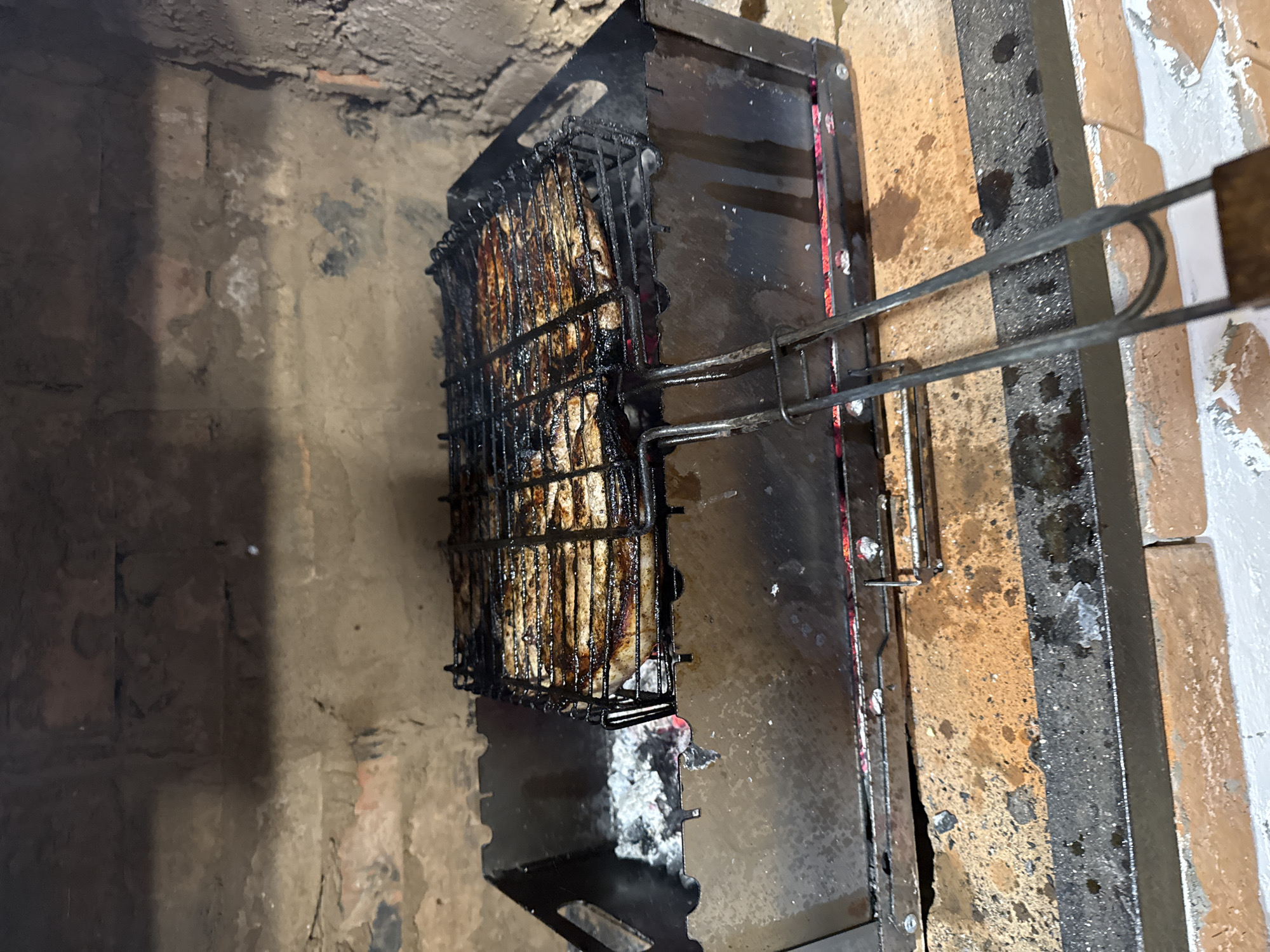 In Gon'ba, Russia, a grill basket filled with seasoned meat is cooking over hot coals inside a brick fireplace. The meat, appearing to be pork ribs or similar cuts, is arranged in rows and is visibly charred in places from the high heat. The grill basket itself is made of dark metal and has two handles for lifting and maneuvering. The fireplace is constructed of rough brickwork, with a metal insert forming the base of the grill. Ash and embers glow beneath the meat, indicating active cooking. The surrounding area appears to be an outdoor or semi-outdoor setting, with a floor made of rough concrete or stone.