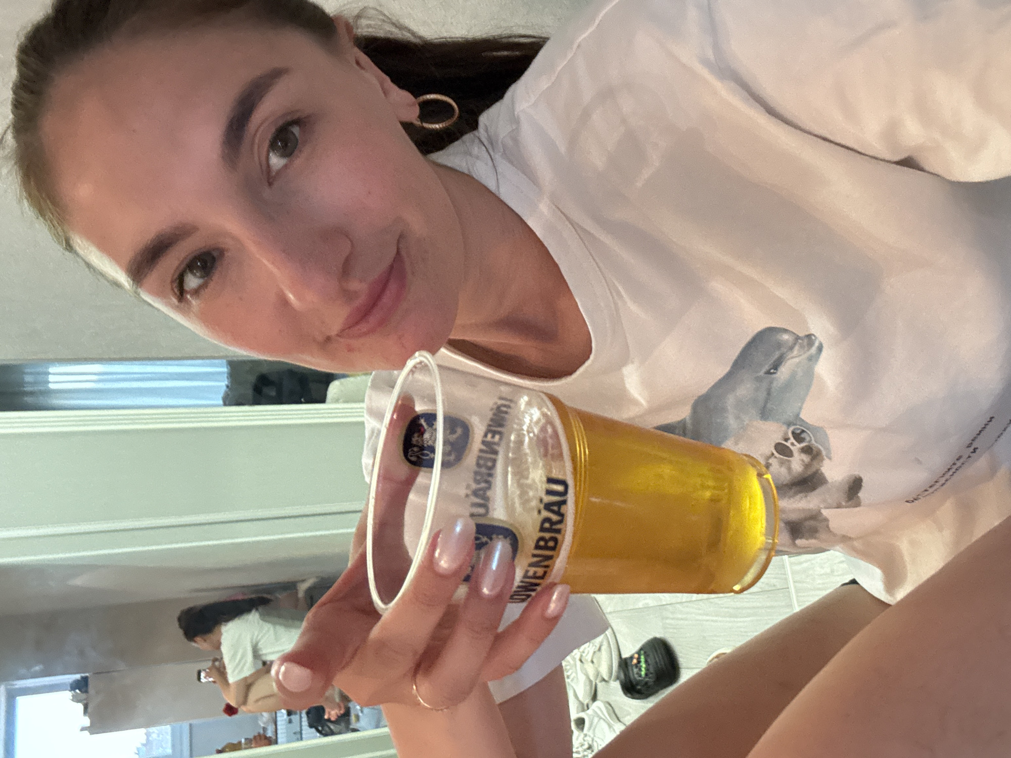 A young woman is holding a plastic cup of beer, specifically Löwenbräu, while taking a selfie. She is wearing a white t-shirt with a dolphin and a cat wearing sunglasses print. She is fair-skinned with dark hair pulled back, and wearing gold hoop earrings. She has a gold ring on her left hand. Her nails are polished in a light iridescent color. The image appears to have been taken in a mirror, as a second woman is visible in the background, seated and looking at a phone. The background shows a shelf with various items and a window, suggesting an indoor setting, possibly a bathroom or living area. While the location in Krasnodar, Russia, is stated, this cannot be confirmed from visual elements in the image.