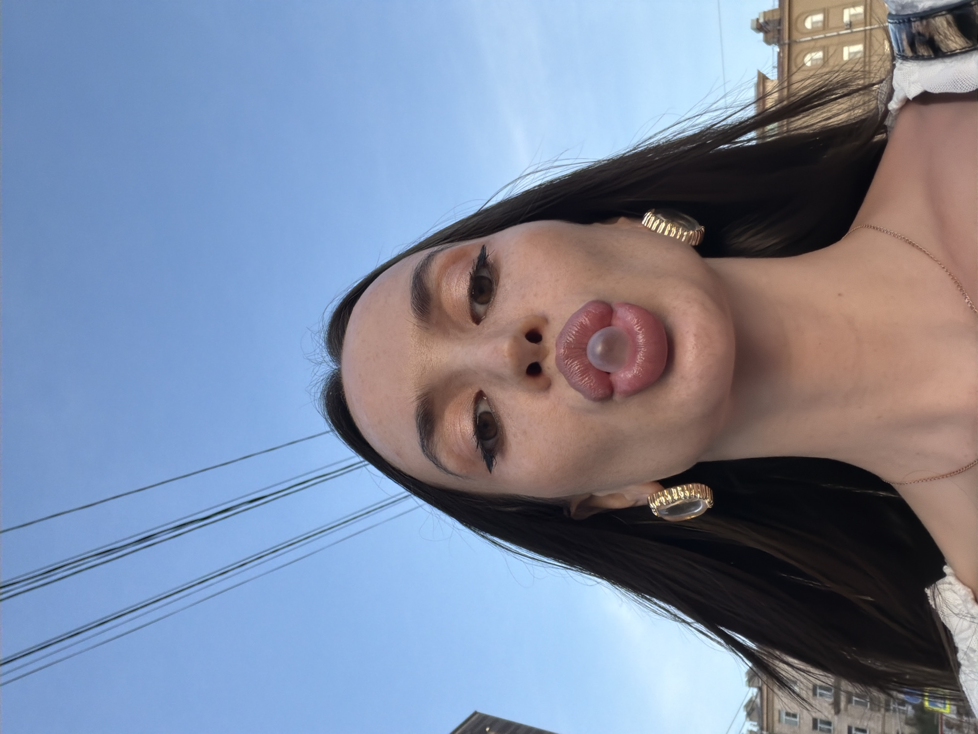An extreme close-up, low-angle shot captures a young woman blowing a bubble with pink gum. She is outdoors, with a clear blue sky and overhead power lines visible. Buildings with windows line the background. The woman has long, dark hair and is wearing large, gold hoop earrings and a delicate gold necklace. Her lips are full and coated with a shimmery gloss, and her eyes are made up with winged eyeliner. The bubble of gum is clear and taut, reflecting light.