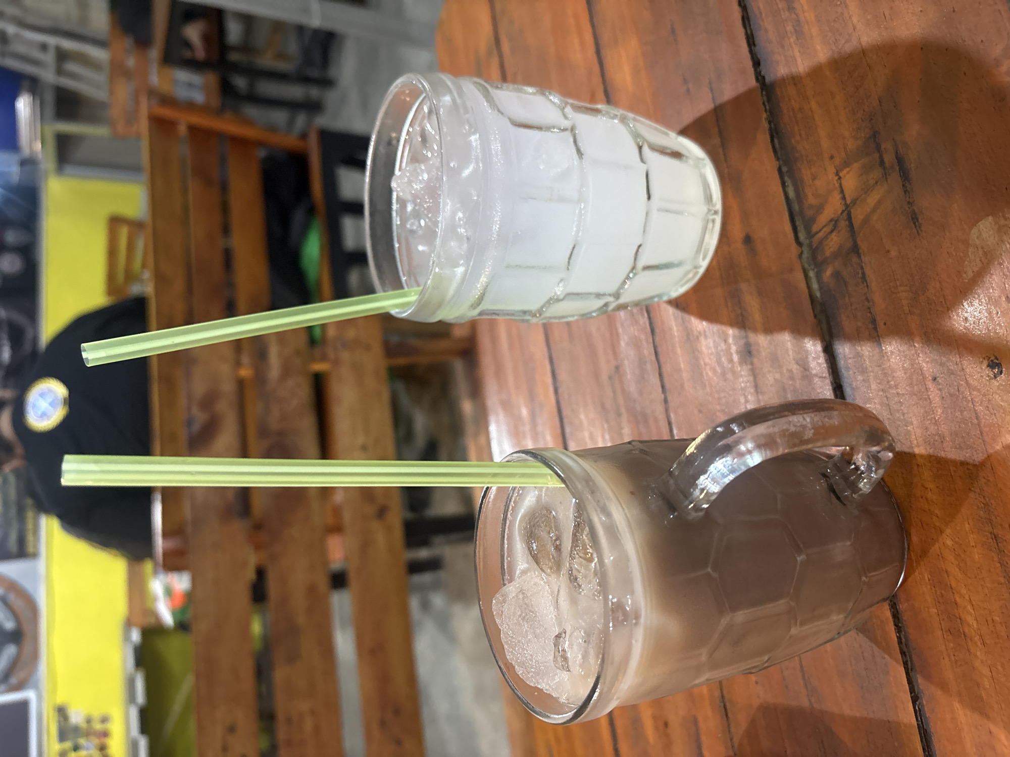 Two beverages with green straws are on a wooden table. The beverage on the left is in a clear mug with a handle and contains a dark brown liquid with ice cubes. The beverage on the right is in a clear glass with a ribbed texture and contains a cloudy white liquid with ice cubes. In the background, a person is seated at a wooden bench. The scene appears to be an outdoor cafe or restaurant.