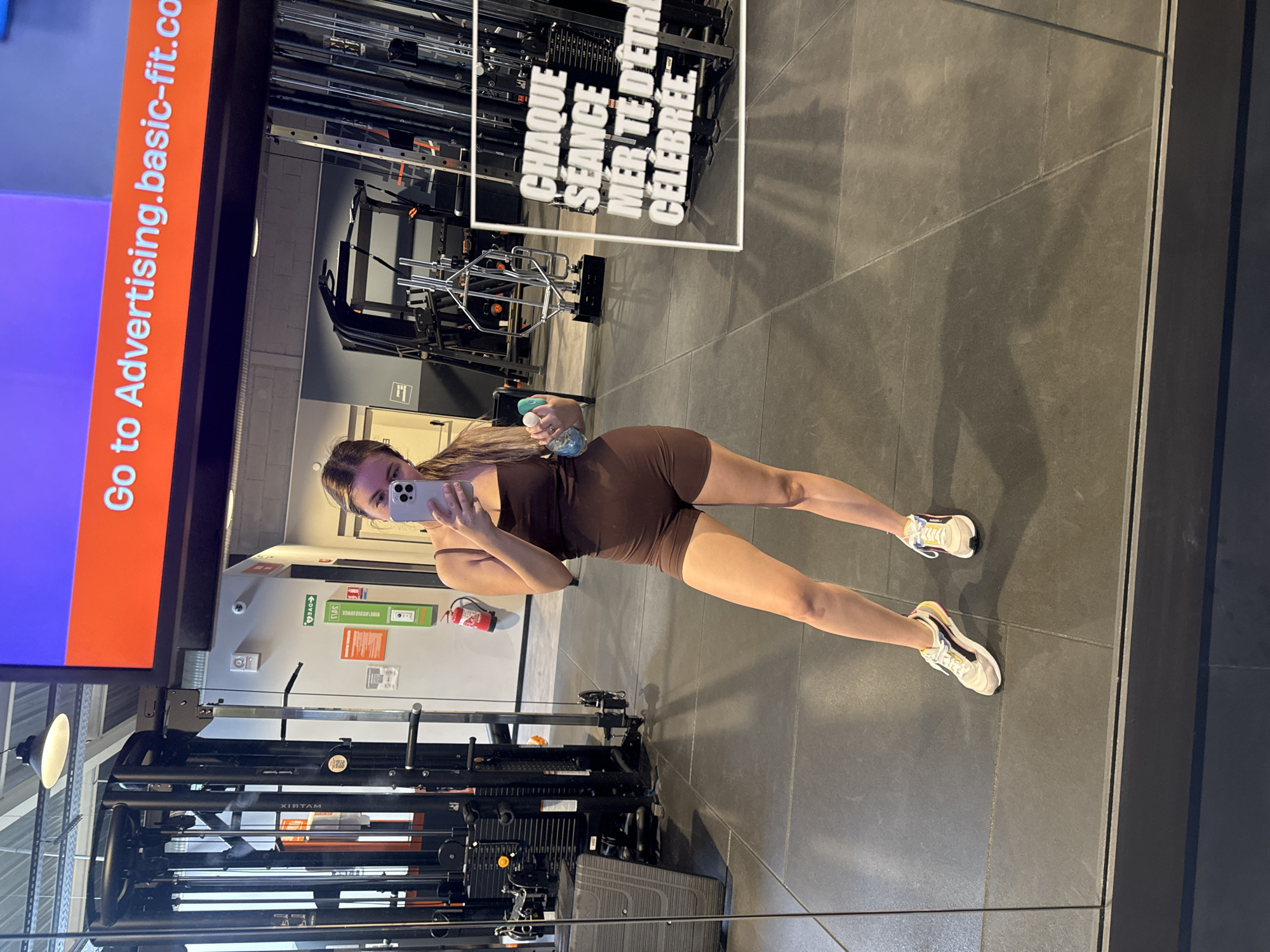 A young woman with long blonde hair, wearing a brown athletic outfit and white sneakers, is taking a selfie in a gym mirror. She is holding a smartphone in her right hand and a water bottle in her left. The gym appears to be modern, with mirrored walls, tiled flooring, and weightlifting equipment visible in the background. A large screen displaying text in French is also present. The lighting is bright, reflecting off the surfaces. The scene is captured from a slightly low angle, focusing on the woman and her reflection.