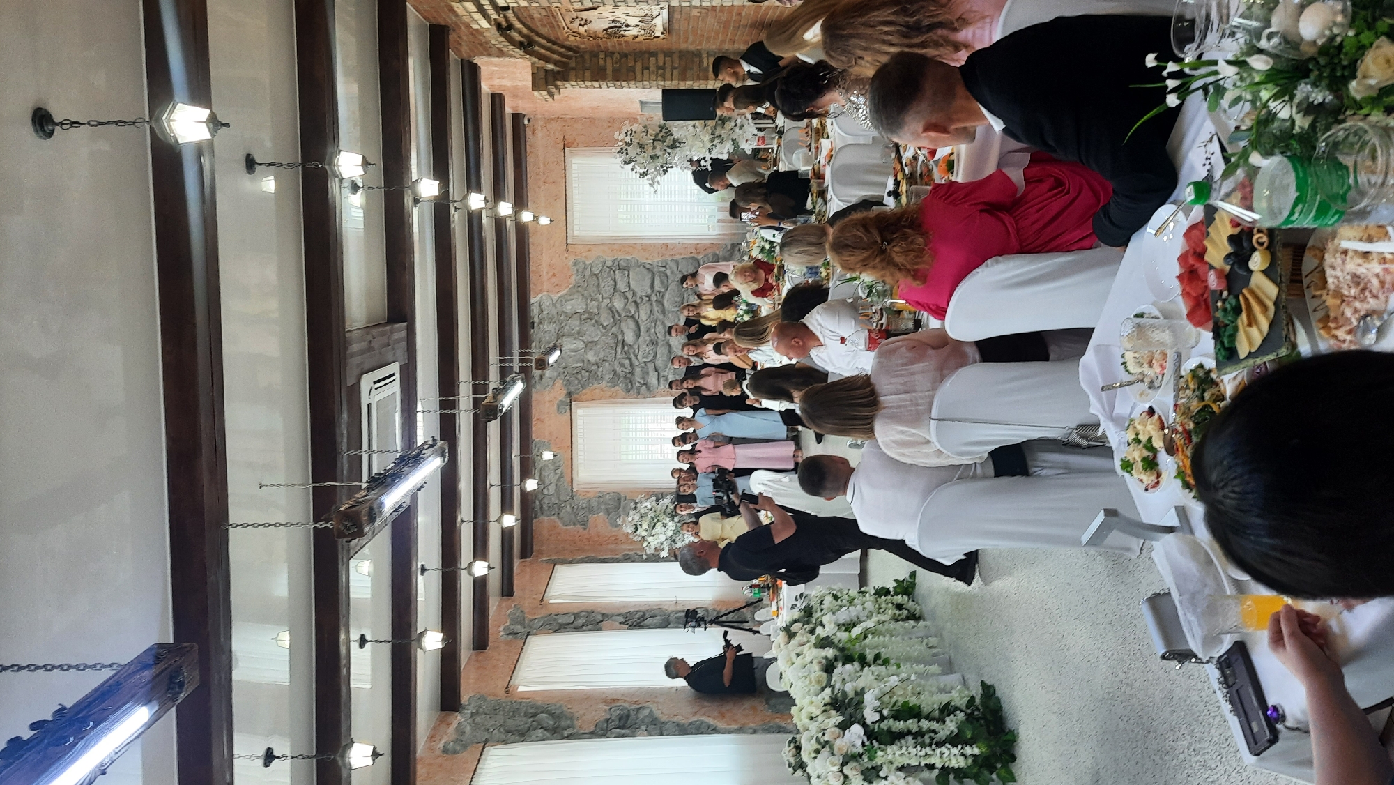 This image captures a formal gathering inside a hall, possibly a wedding reception, in the Zhodziškawski Selski Sovet area of Belarus. A group of people, likely the wedding party or honored guests, stand together at the far end of the hall, dressed in formal attire, with women in pastel-colored dresses and men in suits. In the foreground, guests are seated at tables laden with food and drinks, indicated by plates of various dishes and glasses. Two individuals in the mid-ground appear to be involved in filming or photography, with one holding a camera and the other a tripod. The hall itself features rustic stone walls and wooden beams supporting the ceiling, from which multiple light fixtures are suspended. Large floral arrangements, predominantly white, are placed along the tables and at the end of the hall, adding to the festive atmosphere. The overall scene depicts a celebration in progress, with distinct groups of people engaged in observation and dining.