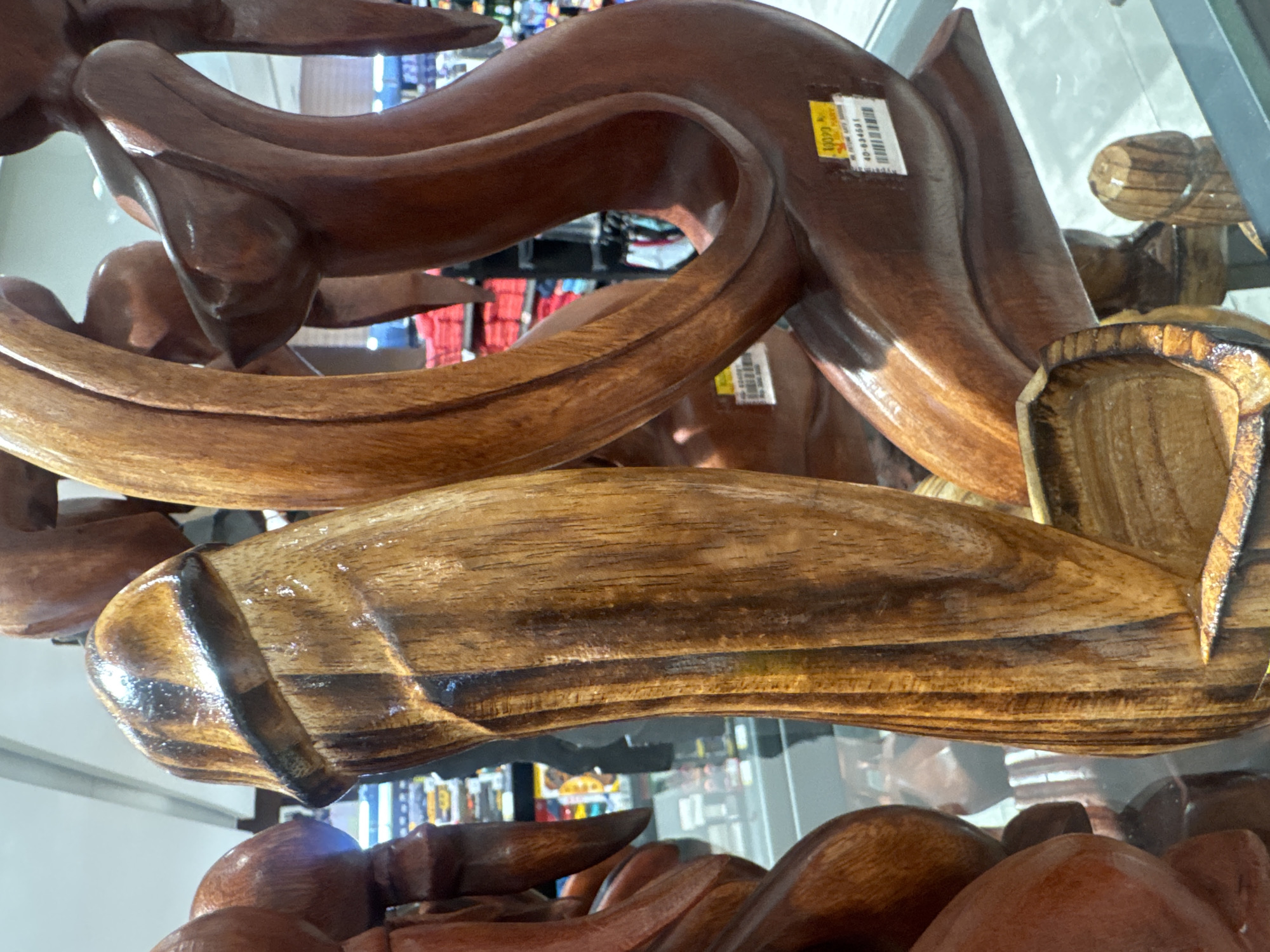 A close-up, eye-level shot focuses on several intricately carved wooden sculptures, presumably displayed for sale in a store. The primary sculpture in the foreground is a phallic-shaped object, carved from a lighter wood with darker striations, showing a polished finish. Behind it, a darker wooden sculpture depicts abstract, flowing human figures, possibly in an embrace. The background shows shelves stocked with various items, suggesting a retail environment. A yellow price tag is visible on the darker sculpture, partially obscuring a barcode and some text in an unfamiliar script. The overall lighting is bright, indicative of indoor store lighting. The scene is located in Kuta, Indonesia.