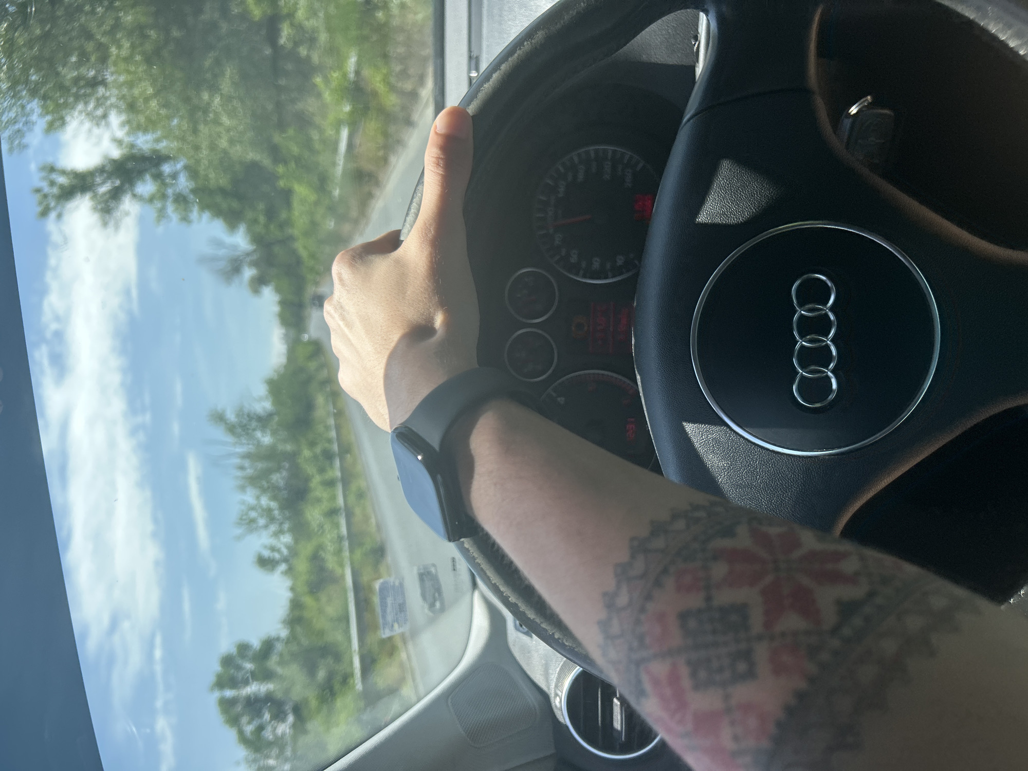 The image provides a first-person perspective from inside a car, showing a person's left arm and hand gripping the top of a steering wheel. A dark-colored smartwatch is visible on the wrist, and the forearm is adorned with a detailed tattoo featuring red and black geometric and floral patterns.
The steering wheel clearly displays the Audi logo, consisting of four interlocking rings. Through the steering wheel, part of the car's instrument cluster is visible, including a speedometer scaled up to 260 km/h and a tachometer reaching 7000 RPM. Various indicator lights and a small digital display showing what appears to be "12:51" or "1251" are also discernible.
Outside the car, through the windshield, a bright daytime scene unfolds. The sky is clear or partly cloudy, and the view shows green trees lining what appears to be a road or highway. Reflections of the sky and foliage are visible on the windshield. The bright lighting suggests it is sunny. The scene depicts someone driving during the day in an area consistent with rural or semi-rural Bulgaria, specifically Mihiltsi, based on the provided context.