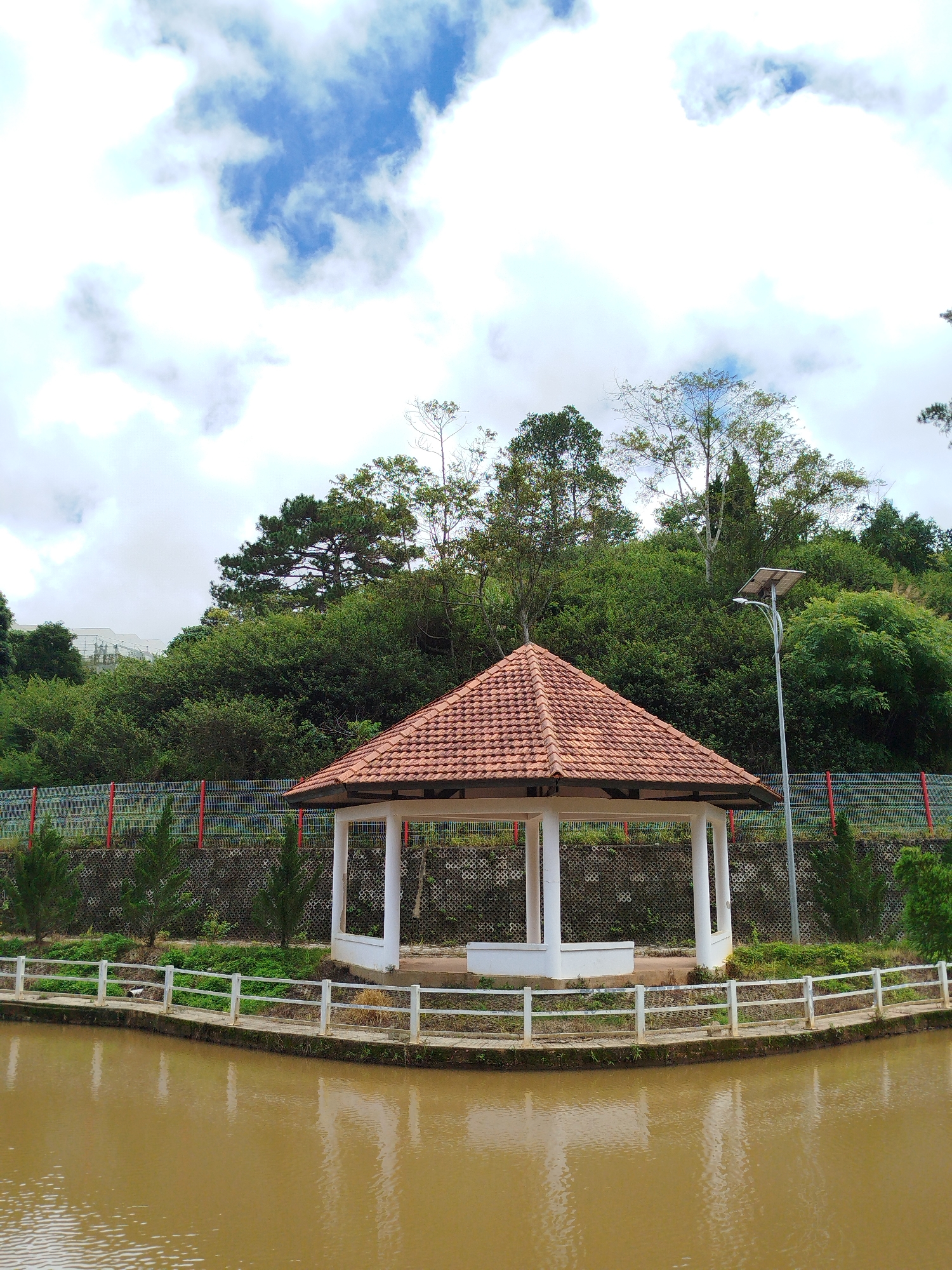 A gazebo with a terracotta tile roof is situated beside a brown, murky body of water in Lạc Dương, Vietnam. The gazebo has a white, open-air structure supported by white pillars, with a low white bench inside. A white railing lines the edge of the water, separating it from a pathway. Beyond the railing, a sloped grassy area is covered with small trees and a concrete retaining wall with a metal fence on top. A solar-powered street light stands to the right of the gazebo. In the background, a dense forest covers a hillside, with some buildings visible on the far left. The sky above is a mix of white clouds and patches of blue.