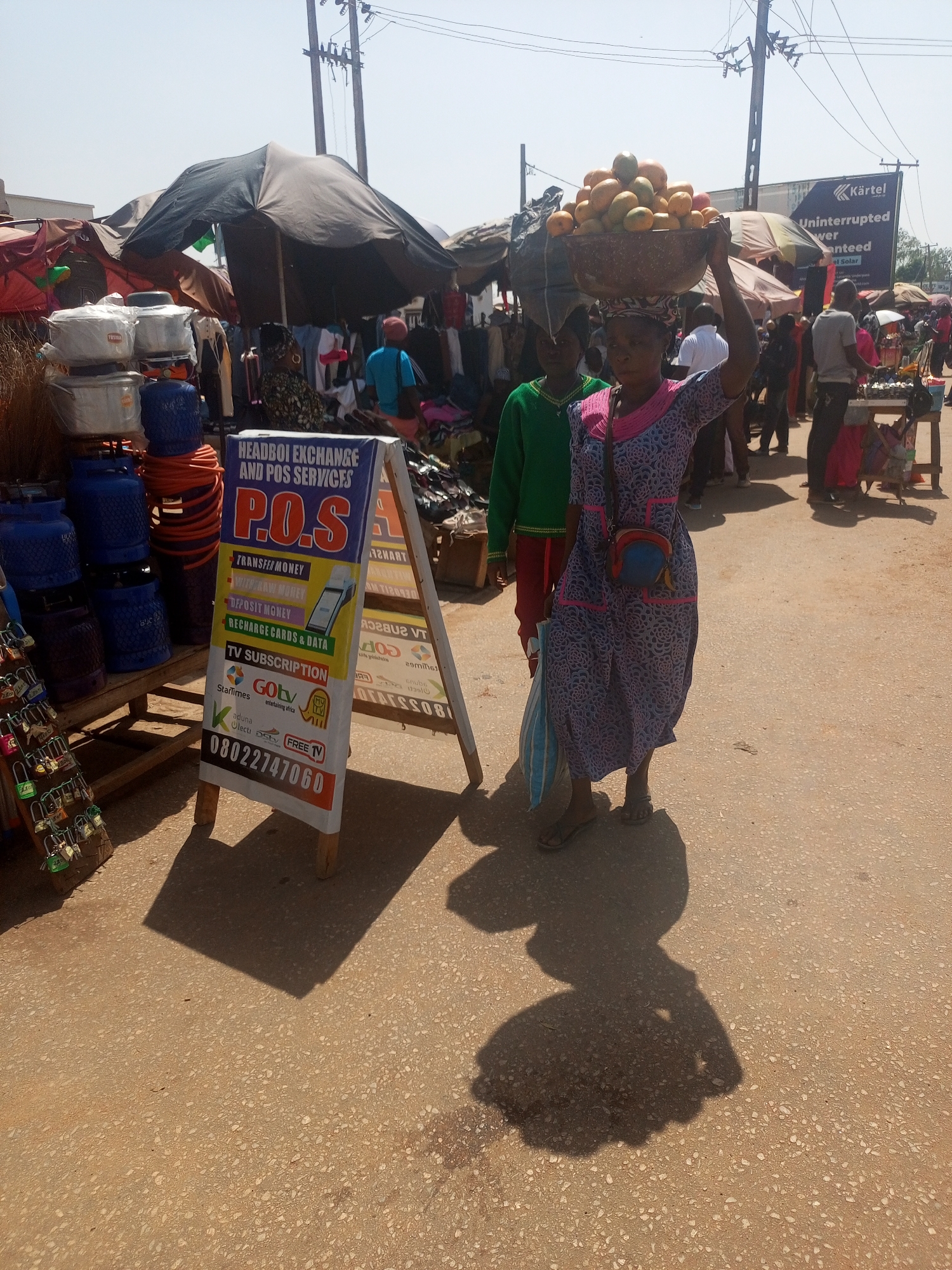 The image depicts a street scene in Sabon Tasha, Nigeria. A woman is carrying a large bowl of mangoes on her head, likely for sale. She is wearing a patterned dress, sandals, and has a small bag slung across her shoulder. A younger person stands behind her, wearing a green shirt and red trousers. To the left is a sign for "HEADBOI EXCHANGE AND POS SERVICES," advertising services like money transfer, TV subscriptions (GoTV, StarTimes, Free TV), and data recharge. The phone number 08022747060 is displayed on the sign. Various merchandise including pots and gas cylinders are stacked on a table next to the sign. The street is busy with other vendors and shoppers under umbrellas and makeshift stalls. A large billboard advertising "Kärtel Uninterrupted Power Guaranteed" is visible in the background. The street surface appears to be paved.