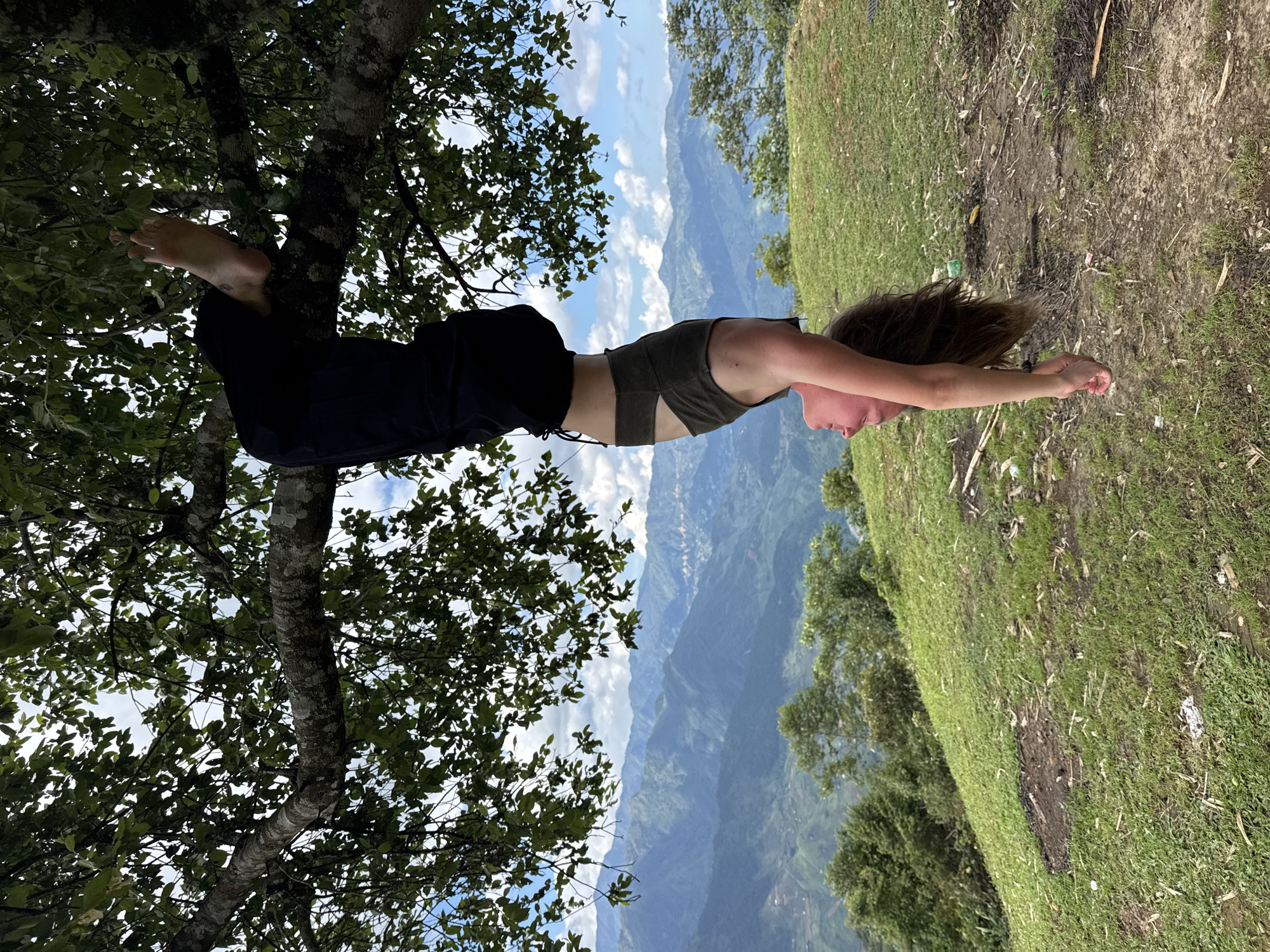 A young adult with light skin and brown hair hangs upside down from a thick, moss-covered tree branch, gripping it with their bare feet. They are wearing a dark olive-green sports bra and loose-fitting black pants with ties at the ankles. Their face is visible, looking towards the ground, with a slight smile and open mouth. Below the person and extending into the background is a vast mountainous landscape, characterized by numerous green, layered peaks receding into a hazy distance under a partly cloudy sky. The immediate foreground shows a grassy hillside with some patches of bare earth and scattered debris, including what appears to be discarded plastic and wood fragments. The scene is located in Tú Lệ, Vietnam.