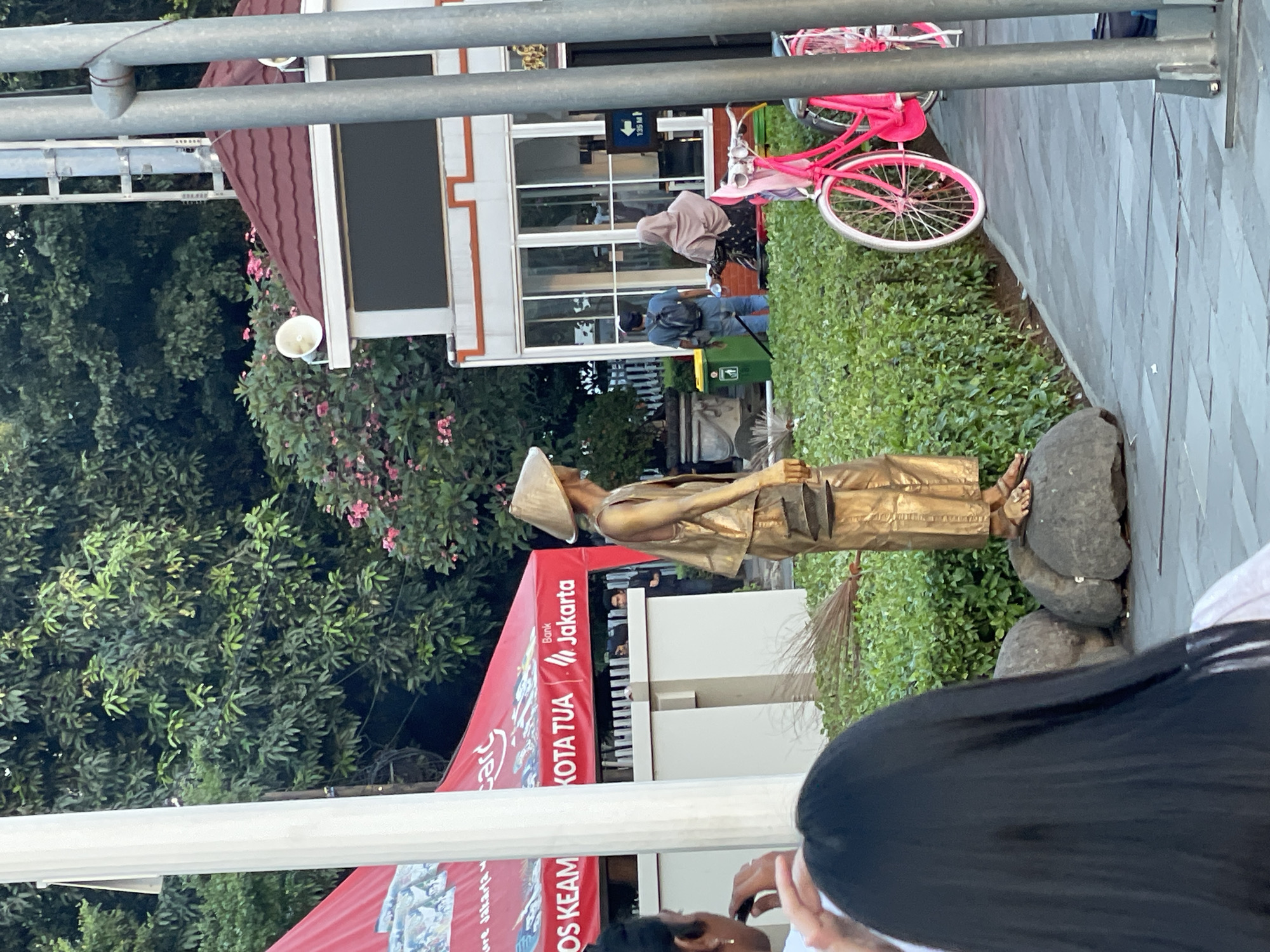 The image depicts a street scene in Jakarta, Indonesia, likely within the Kota Tua area, given the presence of a red tent with "KOTA TUA" branding. In the foreground, a person with long black hair is partially visible, facing away from the camera. The central focus of the image is a living statue, a person adorned in gold paint, wearing a conical hat and traditional clothing, holding a broom. The statue is positioned on large rocks amidst lush green bushes. Behind the statue, a building with a red tiled roof and large glass windows is visible, and a bright pink bicycle is parked to the right. In the background, trees and a red tent further establish the outdoor setting. Several people are seen in the middle ground, including a man with a backpack and another wearing a hijab, suggesting a public space with pedestrian activity. The overall scene suggests a lively urban environment, possibly a tourist attraction or a cultural event.