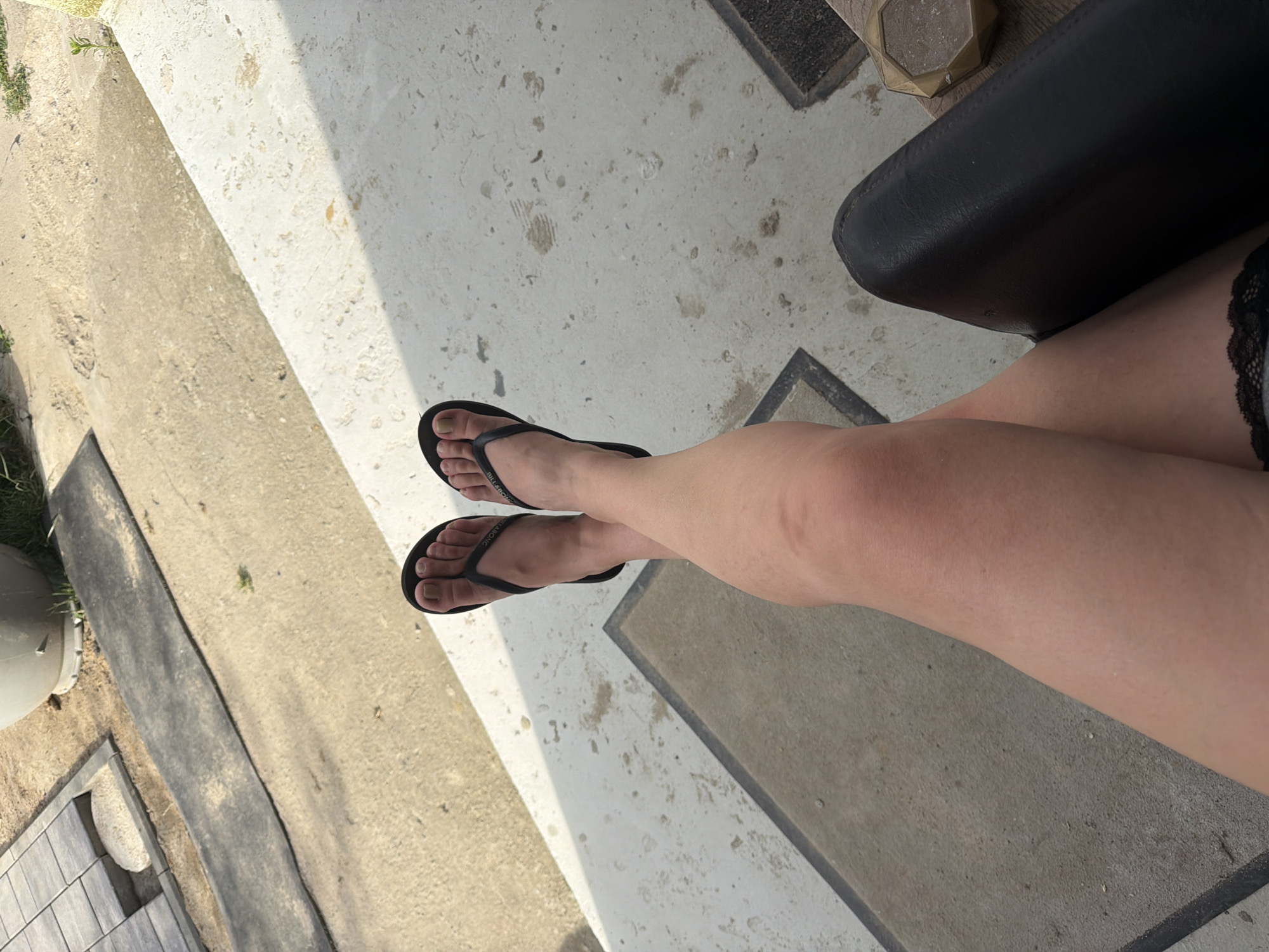 This is a close-up, overhead shot of a person's legs and feet, wearing black flip-flops. The person's legs are crossed and resting on a concrete surface, with a black chair visible to the right. The background shows a patterned paved area with some scattered debris and a large grey planter. The overall lighting suggests an outdoor setting, possibly a patio or balcony, during daylight hours. The image does not contain any elements that would allow for its specific location to be identified as Szczecin, Poland.