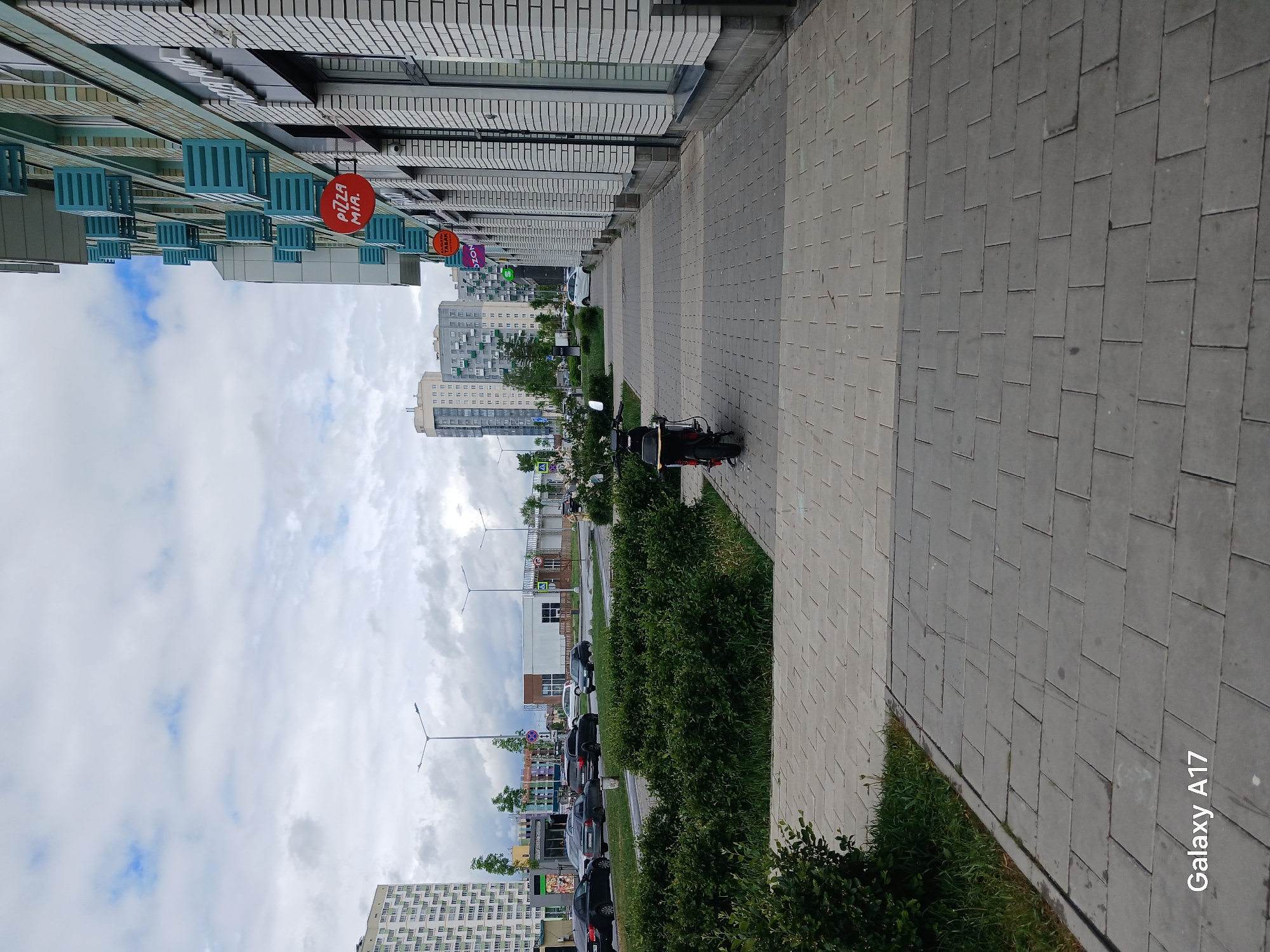 The image shows a street scene in Yekaterinburg, Russia. The camera is oriented along a sidewalk constructed of gray pavers and bordered by a strip of greenery. To the left of the sidewalk are low hedges separating it from a street with parked cars and sparse trees. In the background are apartment buildings of varying heights and architectural styles. Above the buildings is a cloudy sky. Along the building adjacent to the sidewalk, signs are visible, including a "Pizza Mia" sign. There is a motorcycle parked on the sidewalk. A "Galaxy A17" watermark is in the bottom right corner of the frame, indicating the device used to take the photograph.