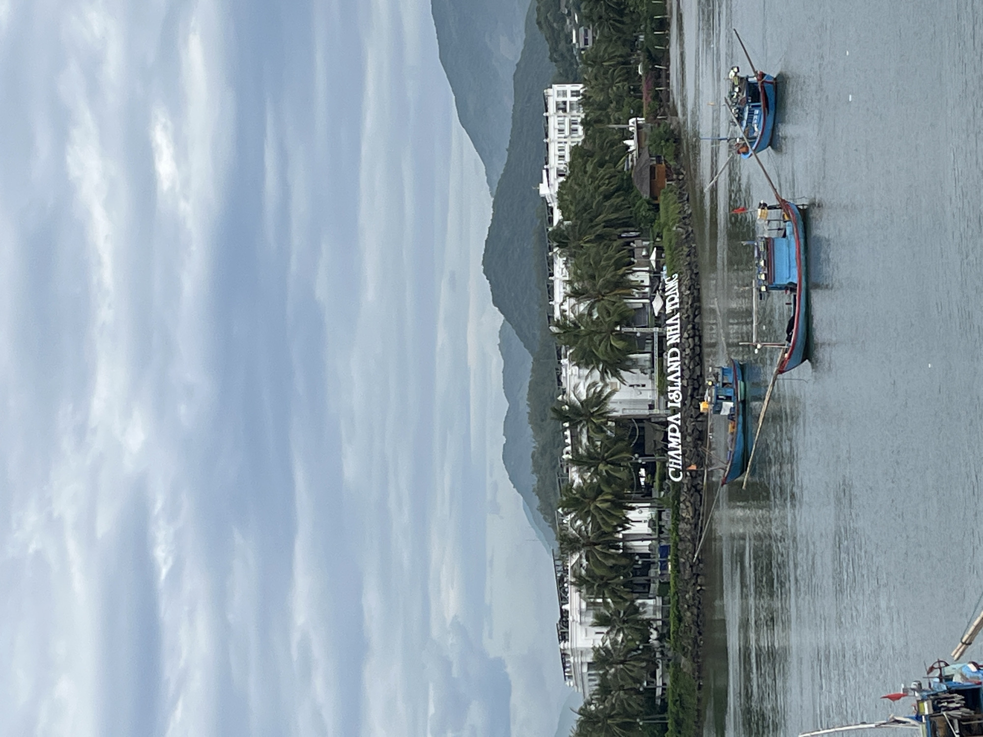 The image captures a scenic view of the Champa Island Nha Trang, a resort located in Nha Trang, Vietnam. In the foreground, a body of calm water is dotted with several blue fishing boats, their fishing rods extended. The middle ground features a long, white, multi-story resort building, its architecture classic and elegant, surrounded by lush green palm trees. Behind the resort, verdant mountains rise, their slopes covered in dense vegetation, indicating a natural landscape. The sky above is overcast with gray clouds, creating a diffused light that casts a soft glow on the entire scene. The text "CHAMPA ISLAND NHA TRANG" is visible in white lettering on the shoreline in front of the resort, further confirming the location. The overall atmosphere is tranquil and picturesque, depicting a typical coastal resort setting.