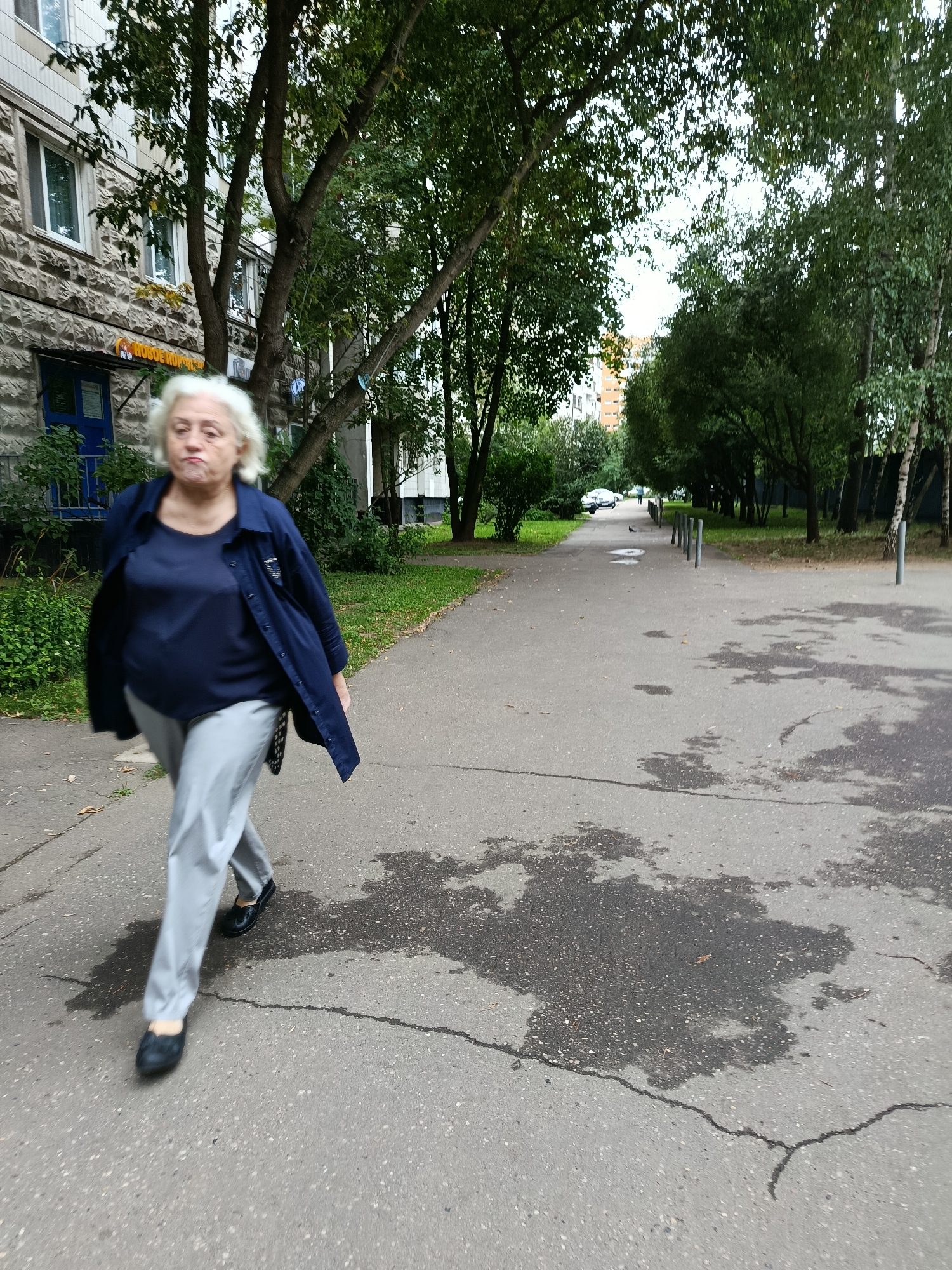 A middle-aged woman with short, white hair walks on a paved sidewalk in front of a building with a rough, grey stone facade. She is wearing a dark blue collared shirt, a dark blue jacket, and light grey pants. She has black shoes on her feet. The sidewalk is wet in patches, suggesting recent rain. Trees with green leaves line the path on the right side of the image, and the building is visible on the left. The environment appears to be an urban setting, likely a residential area in Moscow, Russia, given the architectural style and vegetation.