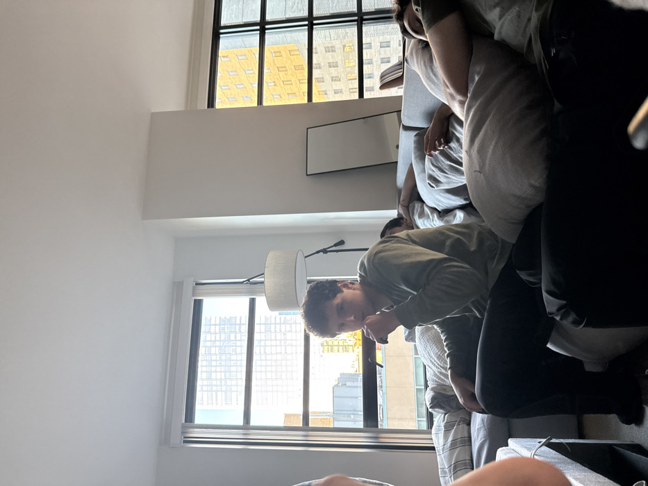 The image depicts a modern, brightly lit apartment interior in a dense urban area, identified as being in Boston, United States. A young adult male with dark curly hair, wearing an olive green long-sleeved shirt and dark pants, sits upright on a grey sectional sofa in the left-center foreground. His right hand is raised to his mouth, possibly holding a small, dark object. He is looking directly towards the camera. To his right, on the same sofa, a second individual is partially obscured, lying down and covered by a light-colored blanket or pillow. Further to the right, a third individual is reclined on the sofa, mostly covered by a grey pillow, resting or sleeping. A fourth person's arm and shoulder are partially visible on the far left of the frame.
The background features large windows revealing multiple high-rise city buildings under bright daylight, including one with a distinct yellowish facade. A modern floor lamp with a white drum shade stands behind the central seated figure. A dark-framed rectangular mirror or artwork leans against a white wall to the right of the lamp. The apartment walls are white, and the visible flooring is dark. The overall scene suggests a casual, relaxed daytime setting.
