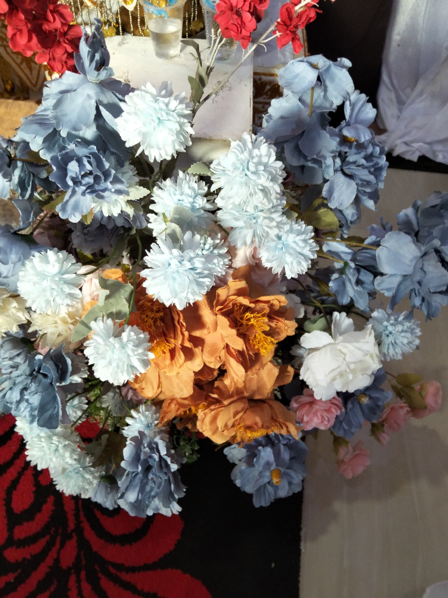 The image shows a close-up, overhead view of a large bouquet of artificial flowers. The bouquet contains flowers in various shades of blue, light blue, white, orange, and pink. Some of the blue flowers are large with multiple layered petals, while others are smaller and round, resembling pom-poms. The orange flowers are large and appear to be peonies, with detailed centers. There are also clusters of white, pom-pom-like flowers and a few pink flowers towards the right side of the bouquet. Green artificial leaves are interspersed throughout. The bouquet is arranged in what appears to be an indoor setting. In the background, a portion of a table with decorative elements and a clear plastic cup with a liquid is visible. The bottom portion of the image shows a dark surface with a red and black patterned design, possibly a rug. There are no people or visible actions taking place. The lighting suggests an indoor environment, and the overall scene is static. The location in Cottrueng, Indonesia, is not visually apparent from the provided image and would require external information.