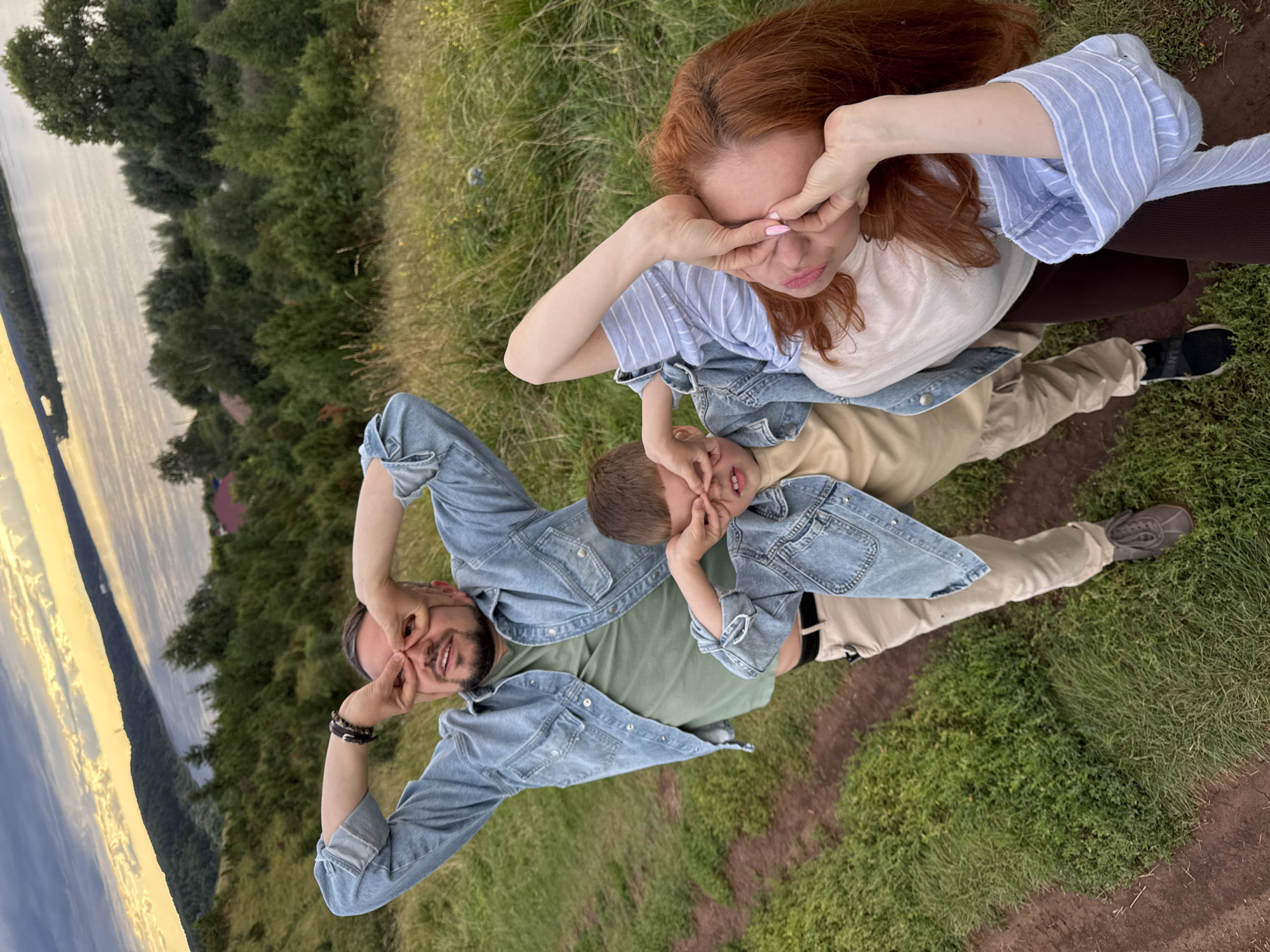 A family of three, a father, mother, and son, are posing for a photo on a grassy hillside overlooking a body of water. They are all wearing denim jackets. The father, in the foreground, is looking at the camera with his hands over his eyes, mimicking binoculars. The son is to his right, also with his hands over his eyes. The mother is on the right, her arm raised to her face with her fingers near her eyes. The landscape behind them features rolling hills covered in trees and a wide river or lake. The sky is overcast with a golden glow near the horizon, suggesting either sunrise or sunset. The terrain is a mix of grass and dirt paths. The scene appears to be in a rural or natural setting.