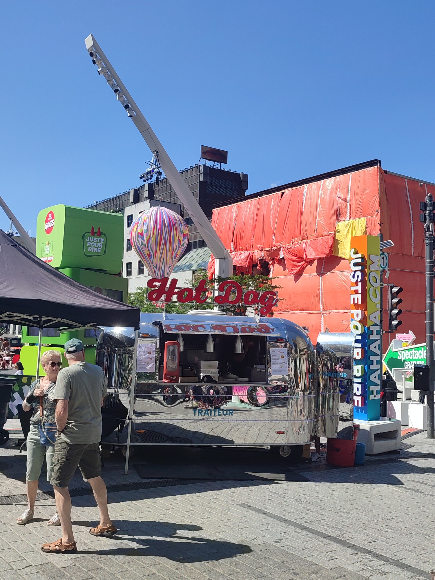 The image captures an outdoor scene in Montreal, Canada, likely during a festival or public event. In the foreground, a shiny, Airstream-style trailer serves as a "Hot Dog" food truck. Two individuals, a woman and a man, are standing near the truck, engaged in conversation. The woman is wearing sunglasses and a patterned top with light-colored pants, while the man is in a t-shirt and cargo shorts.
Behind the food truck, a large building is covered in bright orange tarps, suggesting construction or renovation work. To the left, a bright green, cube-shaped structure with the text "LOL JUSTE POUR RIRE" and a hot air balloon graphic is visible, indicating a connection to the "Juste Pour Rire" comedy festival. A tall, modern light fixture with multiple spotlights extends over the scene from the left.
To the right of the food truck, a colorful vertical sign displays "JUStE POUR RIRE" and "HAHAHA.COM" in bold lettering. Adjacent to this sign is a traffic light and directional signage pointing towards "spectacle GRATUIT," meaning free show. The ground is paved with interlocking bricks, and the sky is clear and blue. The overall atmosphere suggests a lively urban public space.