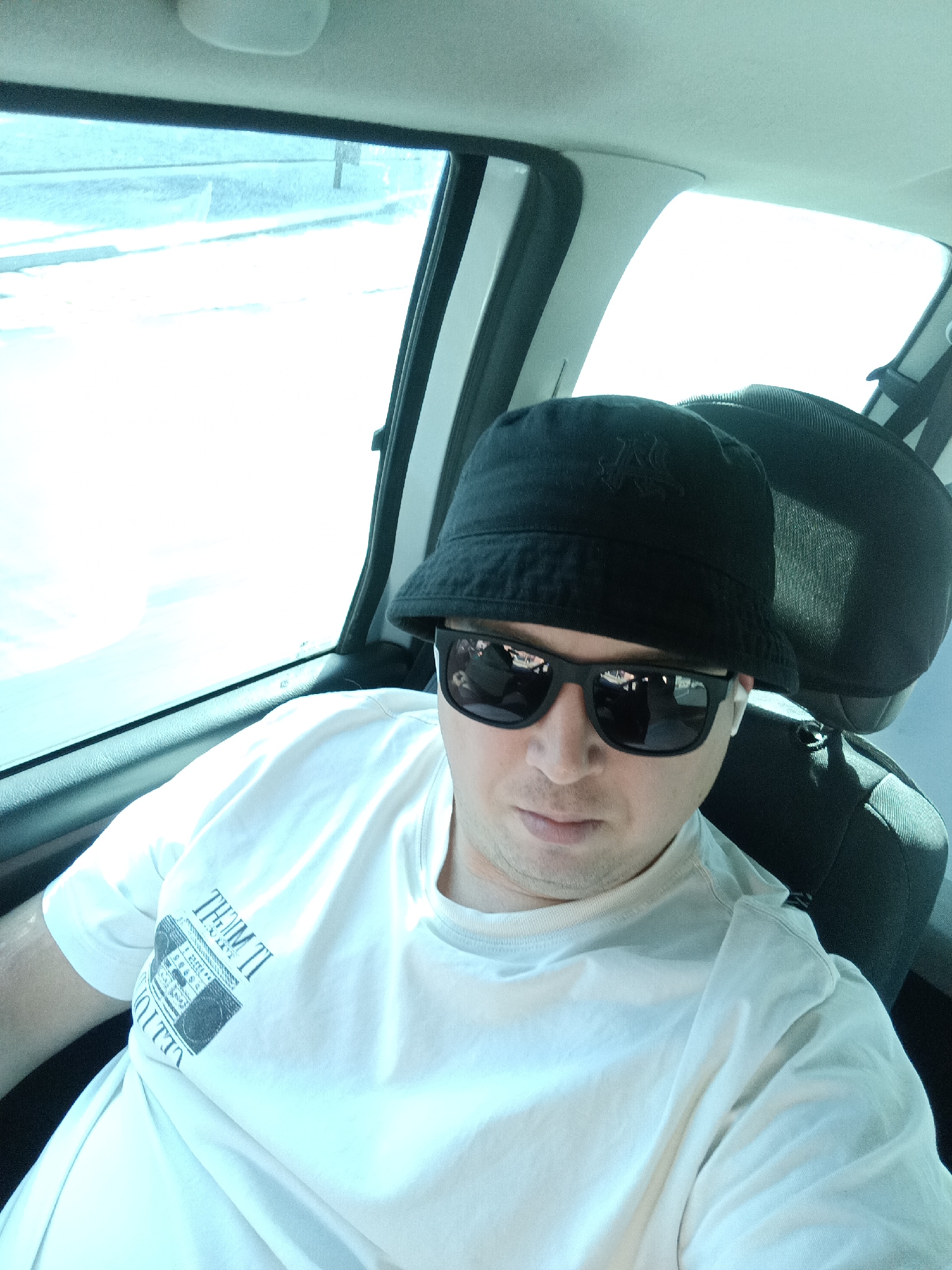 A man is seated inside a vehicle, possibly a car, with his upper body visible. He is wearing a black bucket hat and black sunglasses. His t-shirt is white and has a graphic print on it, partially obscured. The man is looking forward, and his mouth is slightly open. The interior of the vehicle is dimly lit, with bright sunlight filtering in through the windows, creating a strong glare. The background shows the interior of a car seat and the view outside the window, which appears to be a bright, possibly snowy landscape. The specific location of Dalovy, Czechia is not discernable from the image itself.