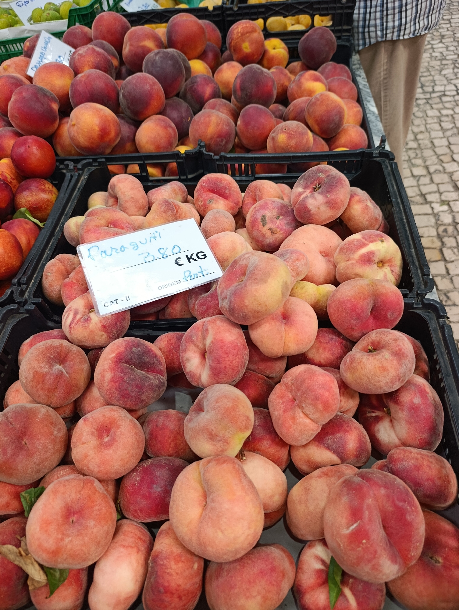 A close-up shot captures a vibrant display of ripe peaches and nectarines, likely at a market stall in Setúbal, Portugal. The fruits, predominantly in shades of red, pink, and yellow, are artfully arranged in black plastic crates, creating a rich tapestry of color and texture. Some peaches are perfectly round, while others exhibit the distinctive flat shape of Saturn peaches, also known as "doughnut peaches." A small white price tag with handwritten text in blue ink is visible, indicating "Paraguai 3.80 € KG" and "CAT. II ORIGEM: Port." This suggests the peaches are of the Paraguai variety, priced at €3.80 per kilogram, with the origin noted as Portugal. The background hints at a cobblestone street and the checkered shirt of a vendor, reinforcing the market setting. The overall impression is one of fresh, local produce ready for sale.