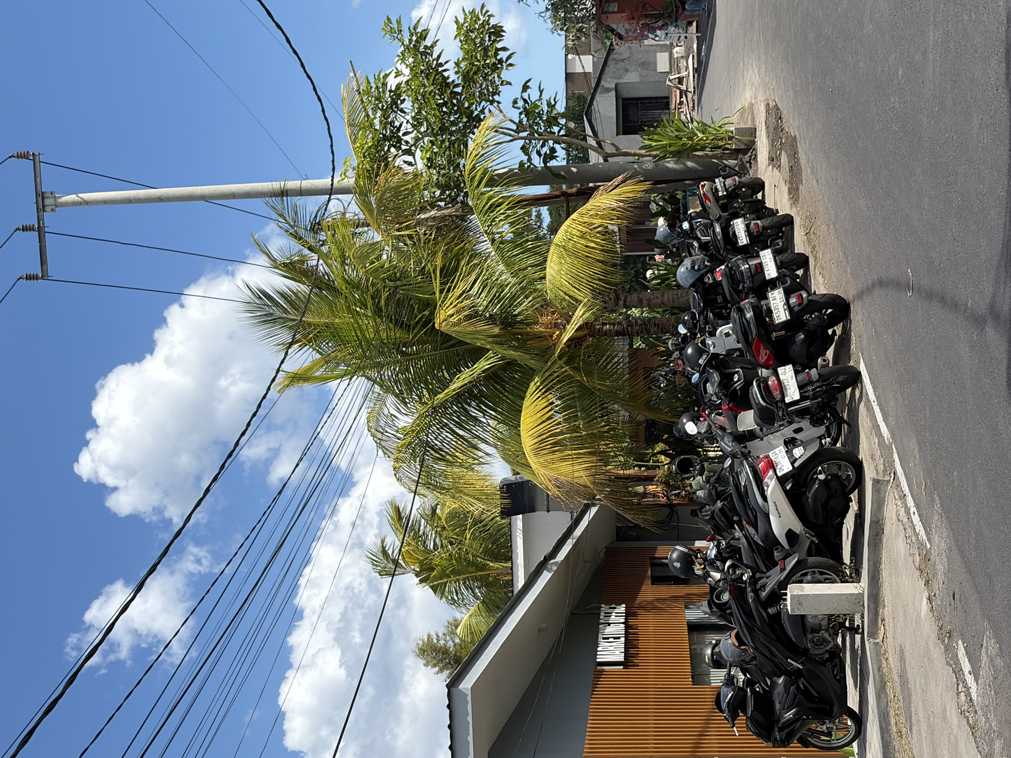 A street scene in Mengwi, Indonesia, features a row of parked scooters along a curb. The scooters are predominantly black and white, with some displaying license plates. Behind the scooters, a modern building with an orange wooden facade is visible, bearing the sign "MARKETING OFFICE". Palm trees with lush green fronds are scattered throughout the scene, some reaching towards a bright blue sky dotted with white clouds. Electrical wires and a utility pole crisscross the sky. Further in the background, there are smaller buildings, one with a grey concrete exterior. The overall impression is a sunny day in a commercial or business area with a tropical feel.