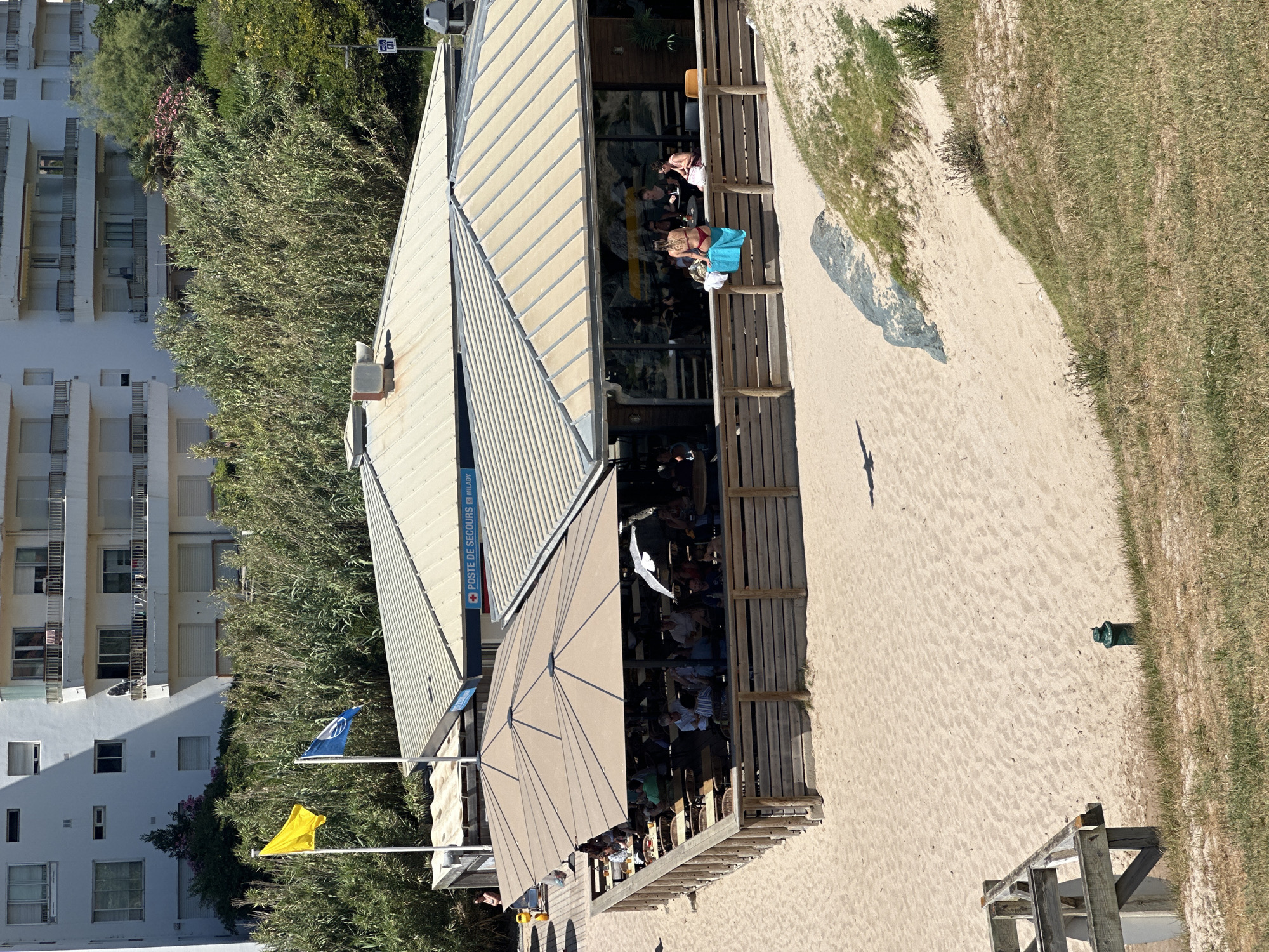 The image shows a beachside restaurant in Biarritz, France, with several people dining under large umbrellas. A building labeled "POSTE DE SECOURS MILADY" is visible, indicating a lifeguard station. Two flags, one yellow and one blue with white markings, fly in front of dense vegetation. The restaurant has a wooden fence surrounding its outdoor seating area, which is on the sand. People are seen sitting at tables, some conversing and others eating. In the foreground, the sandy beach slopes up to a grassy dune.