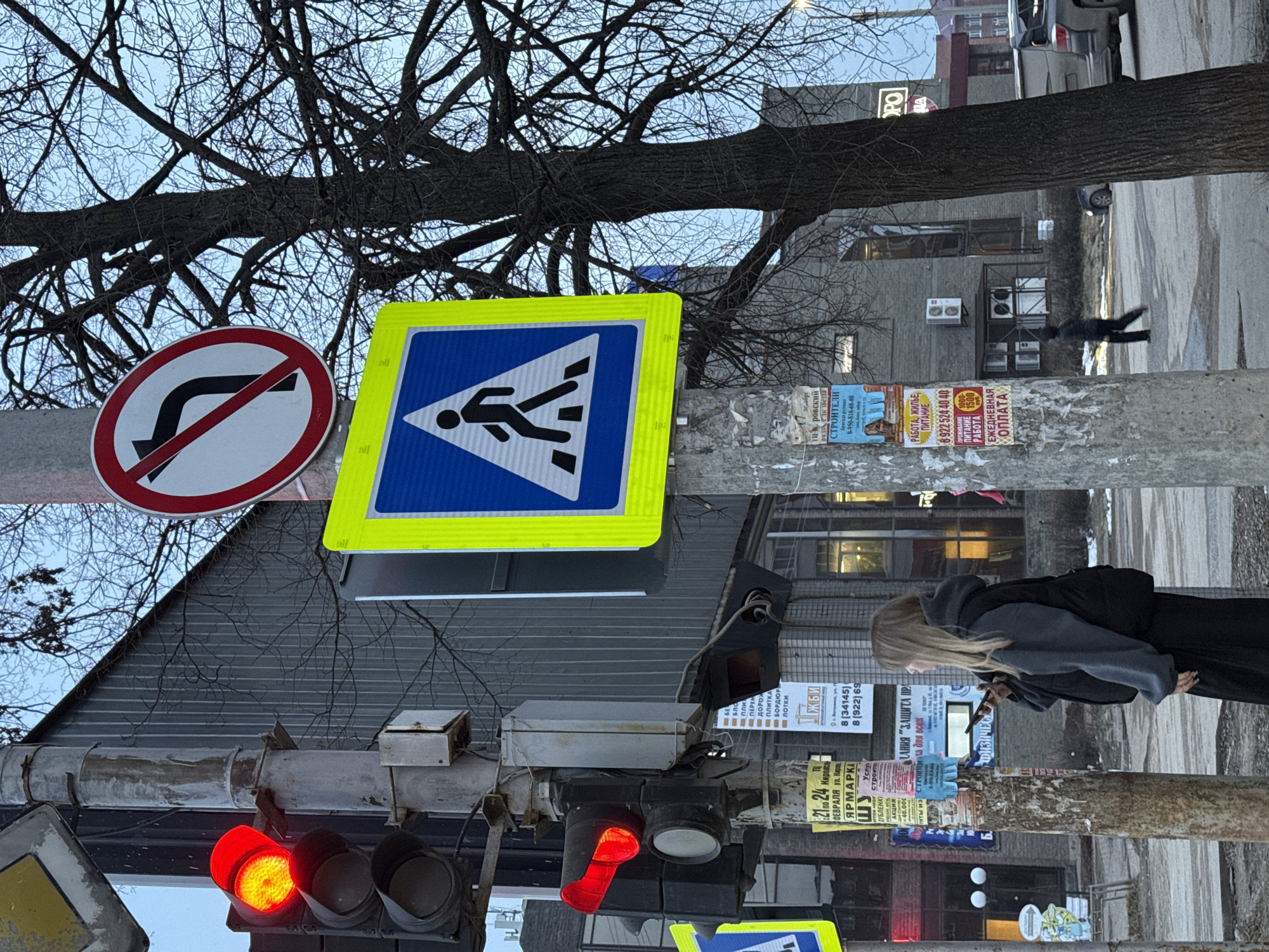 The image depicts a street scene in Votkinsk, Russia, featuring a pedestrian crossing. Traffic lights are visible, with both red and red-and-yellow signals lit. Street signs indicate a pedestrian crossing and a no-right-turn restriction. A utility pole covered in advertisements is present. A woman walks on the sidewalk, seemingly using her phone. In the background, buildings can be seen, and a tree with bare branches overhangs the scene. A person is crossing the street in the background. The text on the advertisements includes contact numbers and promotions for various services.