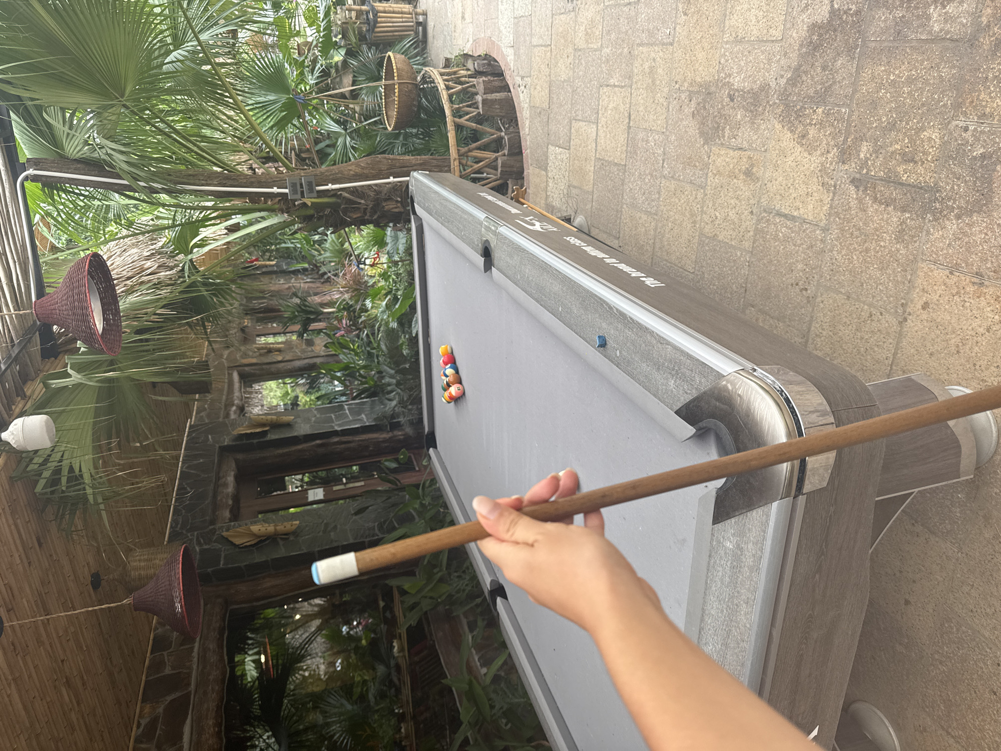 An outdoor pool table is set up on a stone patio in what appears to be a tropical or semi-tropical environment. The pool table is gray with a set of colorful billiard balls racked at one end. A person's hand is visible in the foreground, holding a pool cue, as if about to play. Surrounding the patio are lush green palm trees and other tropical foliage, suggesting a resort or recreational area. In the background, there's a building with a thatched roof and stone accents, further reinforcing a tropical setting. The lighting indicates it is daytime, possibly overcast. The location, Nam Hoa Lư Ward, Vietnam, suggests this could be a leisure area within that region.