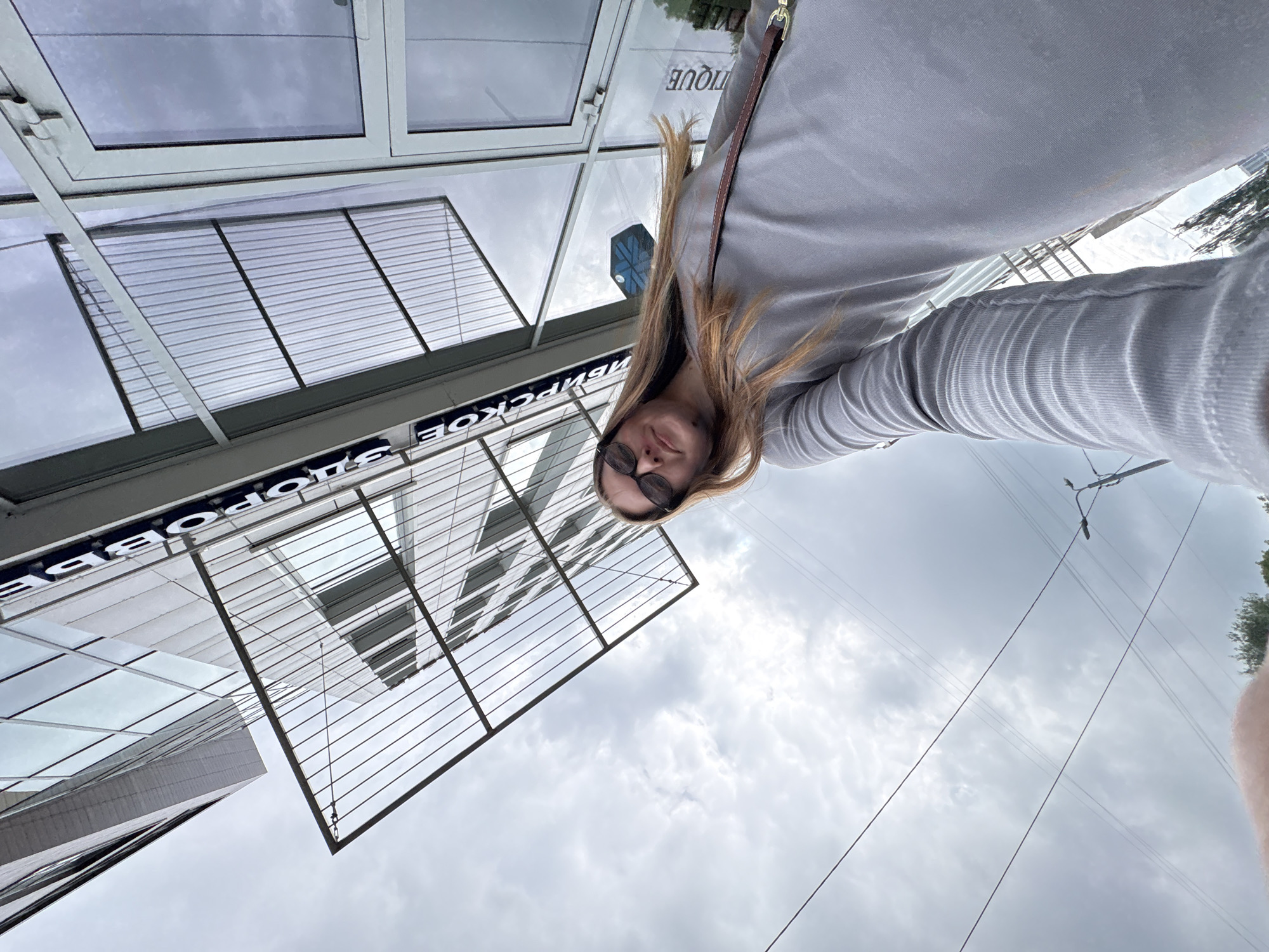 A low-angle, wide shot captures a young woman with long blonde hair and sunglasses, wearing a grey long-sleeved shirt, posing for a selfie in front of a modern building. The building, located in the city of Perm, Russia, features a glass facade and a prominent metal canopy over what appears to be an entrance. Text on the building, partially visible and in Cyrillic, reads "ВИРХОВСКОЕ" and "BOUTIQUE". The sky is overcast with grey clouds, and utility wires are visible crisscrossing the frame, along with a streetlamp on the right side. The perspective is from below the woman, looking up towards her and the building.