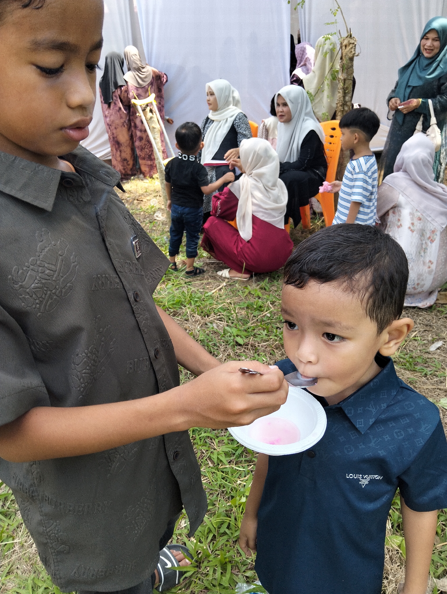 In Pulonga, Indonesia, a young boy in a dark blue Louis Vuitton polo shirt is being fed a spoonful of pink liquid from a white bowl held by an older boy wearing a grey, patterned shirt. The scene appears to be outdoors, possibly at a gathering, with other people and a white tent in the background. Some individuals are wearing headscarves, suggesting a cultural event. The ground is covered in grass and dry leaves.