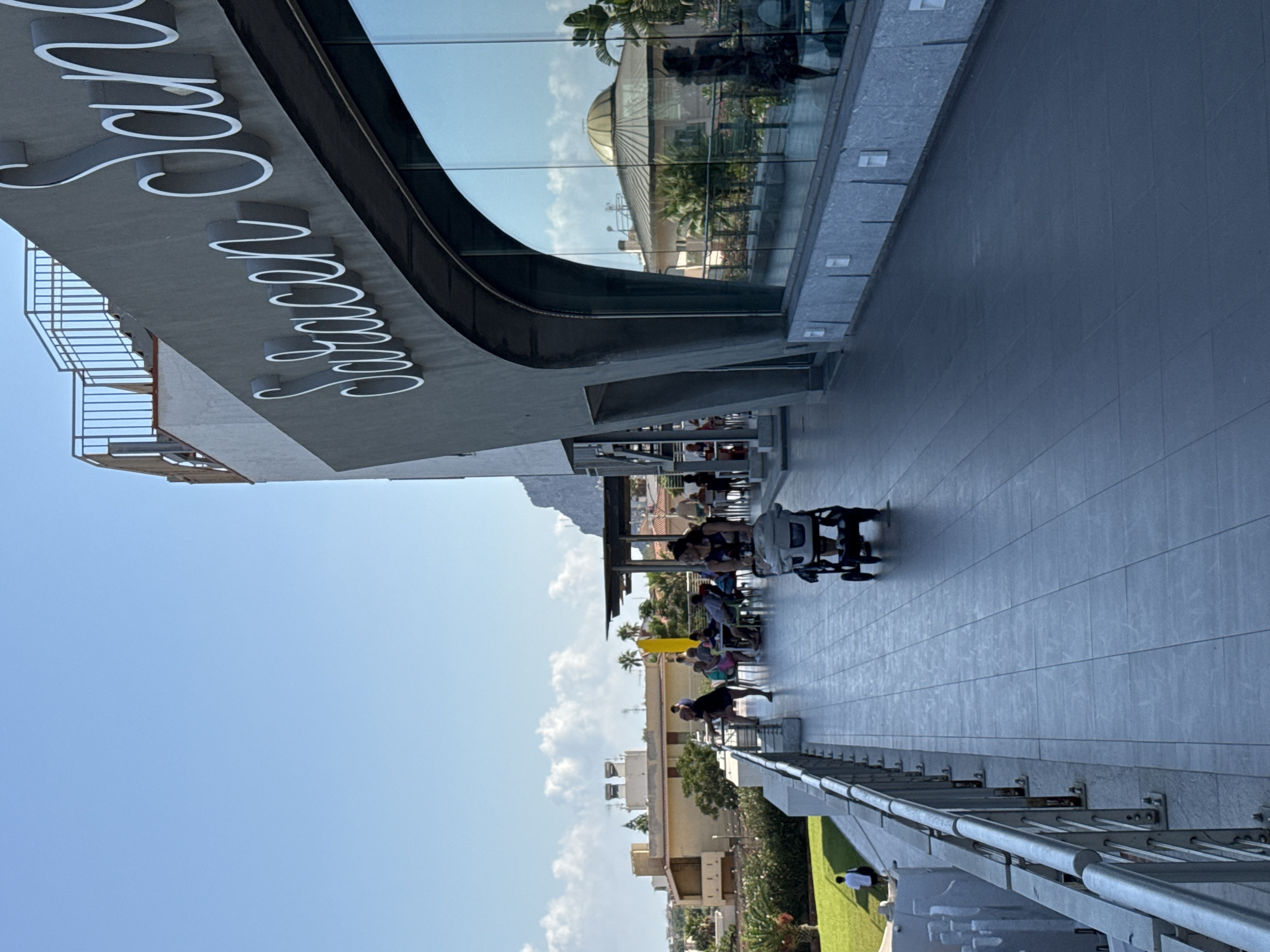 The image shows an outdoor walkway in front of a building with a large glass facade. The building has the name "Saracen" in large, white, raised lettering. The walkway is paved with grey tiles and has a metal railing along its edge. Several people are visible on the walkway and at tables set up outside a restaurant area. One person is pushing a stroller. In the background, there are buildings, trees, and a clear blue sky with some clouds. The overall scene suggests a resort or hotel setting. The text "Saracen" is a reference to the Saracen Sands hotel in Capaci, Italy.
