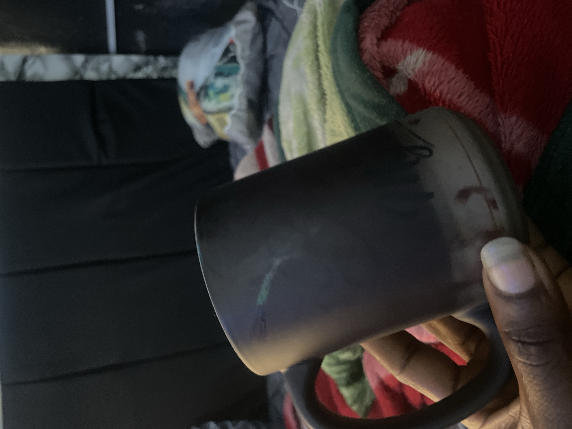 A dark-colored mug is held by a person's left hand, with their thumb resting on the mug's rim and fingers curved around the handle. The mug is dark, with some lighter markings and a slight gradient towards the bottom. In the background, a dark, padded headboard is visible, partially obscured by a patterned throw pillow. Below the headboard, several blankets are spread out, including one with green and yellow stripes and another with red and white stripes. The scene suggests an indoor setting, possibly a bedroom or living room. The specific location within New Stadium, Nigeria, cannot be determined from the image alone.