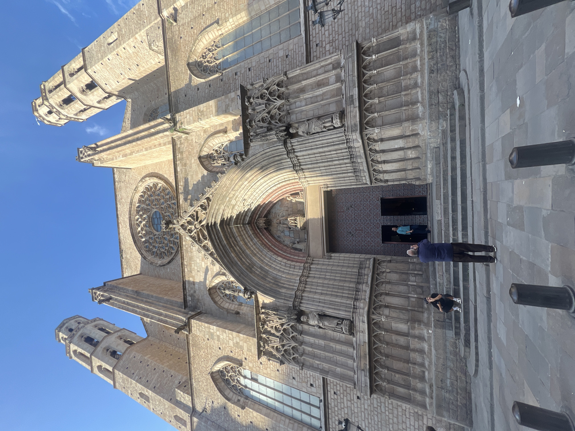 The image displays the impressive Gothic facade of a historic stone building, likely a cathedral or church, under a clear, bright blue sky. The architecture is characterized by intricate stone carvings, pointed arches, and a large rose window dominating the center above the main entrance. Two distinct bell towers rise towards the sky, one on the left and another on the right, both featuring multiple open arches.
The main entrance is a prominent feature, deeply recessed with a grand arched doorway adorned with statues and elaborate decorative work. Sunlight casts sharp shadows, highlighting the architectural details and suggesting a sunny day, likely late morning or early afternoon.
In the foreground, on the stone steps leading up to the entrance, two individuals are visible. One person, wearing a dark blue top and dark trousers, stands facing the building, seemingly engrossed in viewing or photographing the structure. Another person, dressed in dark clothing and light sneakers, crouches lower on the steps, also appearing to be taking a photo or closely examining the building. A third person is partially visible further inside the entrance. Several dark, cylindrical bollards are present on the paved ground near the steps.
The scene depicts a moment of sightseeing or appreciation of historical architecture, consistent with a tourist location in a city like Barcelona, Spain, given the architectural style and provided context. No visible text is discernible in the image.