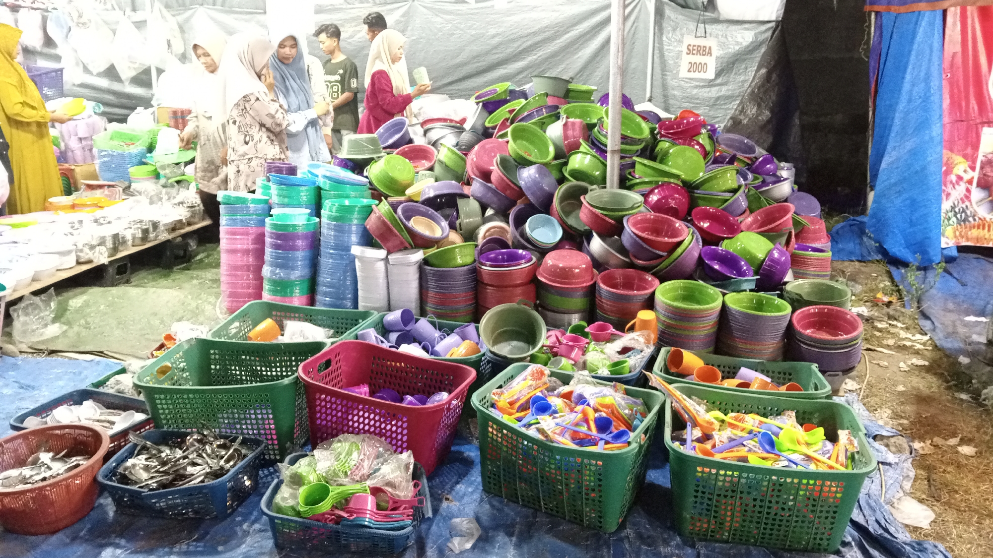 In Cotgadong, Indonesia, an outdoor market stall displays a vast assortment of colorful plastic household items. Stacks of bowls and containers in various shades of pink, purple, green, and blue are piled high, some neatly stacked and others in disarray. Several green plastic baskets are filled with these items, alongside smaller containers holding cutlery and utensils. In the background, several individuals, including women wearing headscarves and men, are browsing the merchandise or attending the stall. A tarp-covered structure forms the backdrop of the stall, with a sign in Indonesian reading "SERBA 2000." The ground is covered with a blue tarp, and surrounding the stall are more goods and debris, indicating a bustling marketplace environment.