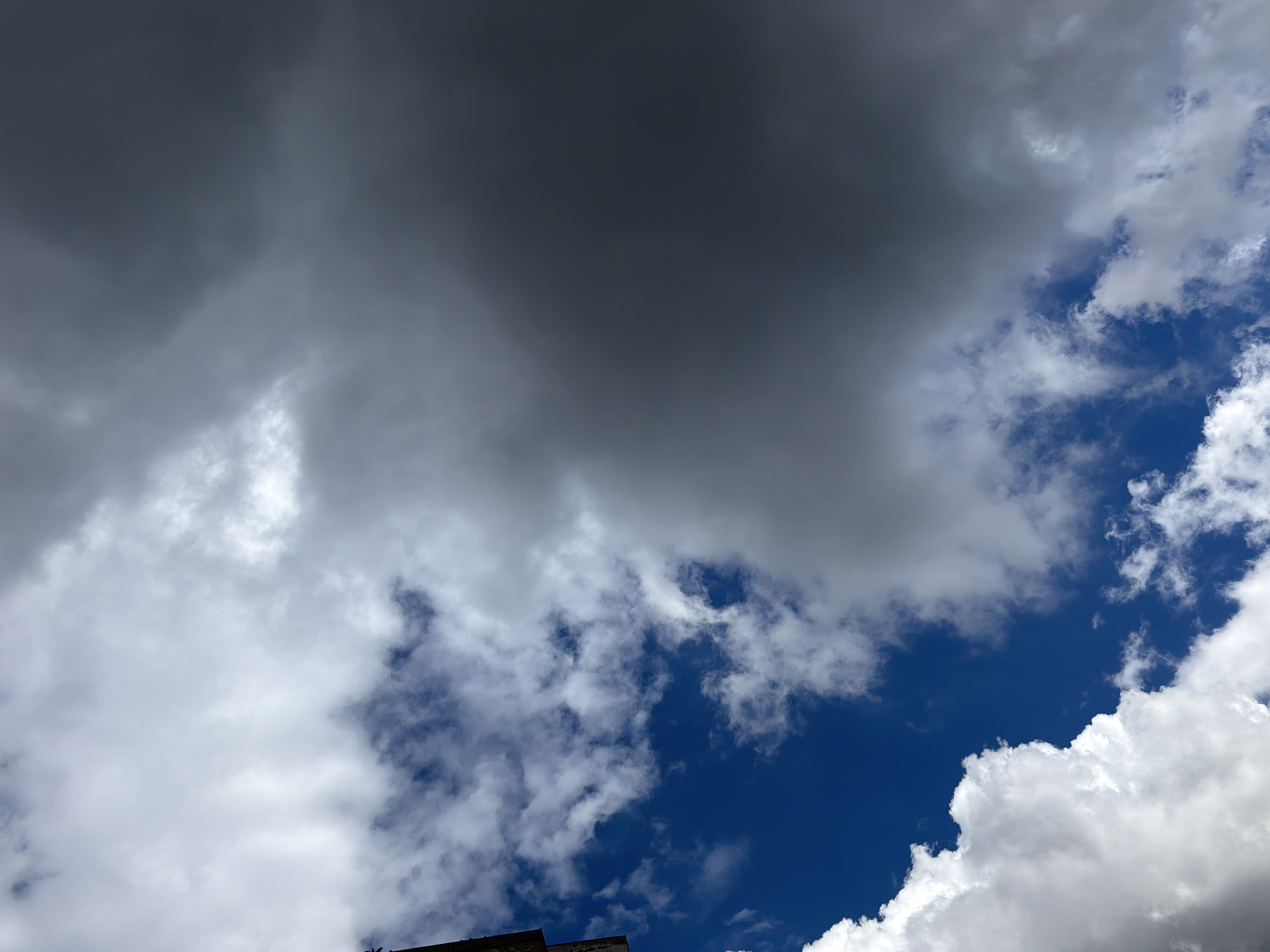 The image displays a dynamic sky scene in Sofia, Bulgaria, characterized by a striking contrast between dark, heavy cloud formations and bright, fluffy white clouds, interspersed with patches of clear blue sky. The upper left quadrant of the frame is dominated by a large expanse of dark grey, dense clouds, suggesting overcast conditions or impending weather change. This darker cloud mass gradually gives way to lighter, more scattered white cumulus clouds towards the right and bottom of the image. Vibrant, deep blue sky is visible in several areas, particularly prominent towards the bottom right and middle sections, creating a sense of depth and openness. At the very bottom edge of the frame, a small, dark silhouette of what appears to be the top of a building or urban structure is barely visible. The scene captures a daytime sky with varied weather, showcasing the interplay of light and shadow across different cloud types. No specific activity, event, people, or visible text are present in the image.
