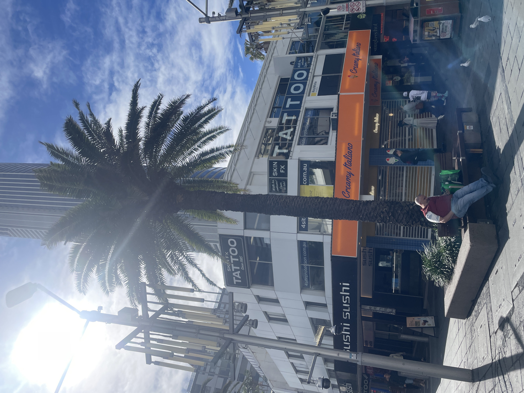 In Surfers Paradise, Australia, a sun-drenched street scene unfolds, dominated by a tall palm tree casting a shadow across the sidewalk. Above, a modern building displays prominent signage for "SKIN FX TATTOO Since 1985" and "4siteprotection.com.au." To the left, another sign reads "sushi sushi." Below, an orange awning stretches across the storefronts of "Creamy Italiano," an ice cream parlor. Several individuals are present: a man in a red vest and blue jeans sits on a bench, seemingly engrossed in his phone, while other people walk by or stand in small groups near the establishments. Pigeons peck at the ground in the foreground. The environment is urban, with paved sidewalks, streetlights, and other buildings visible in the background, under a bright blue sky with scattered clouds.