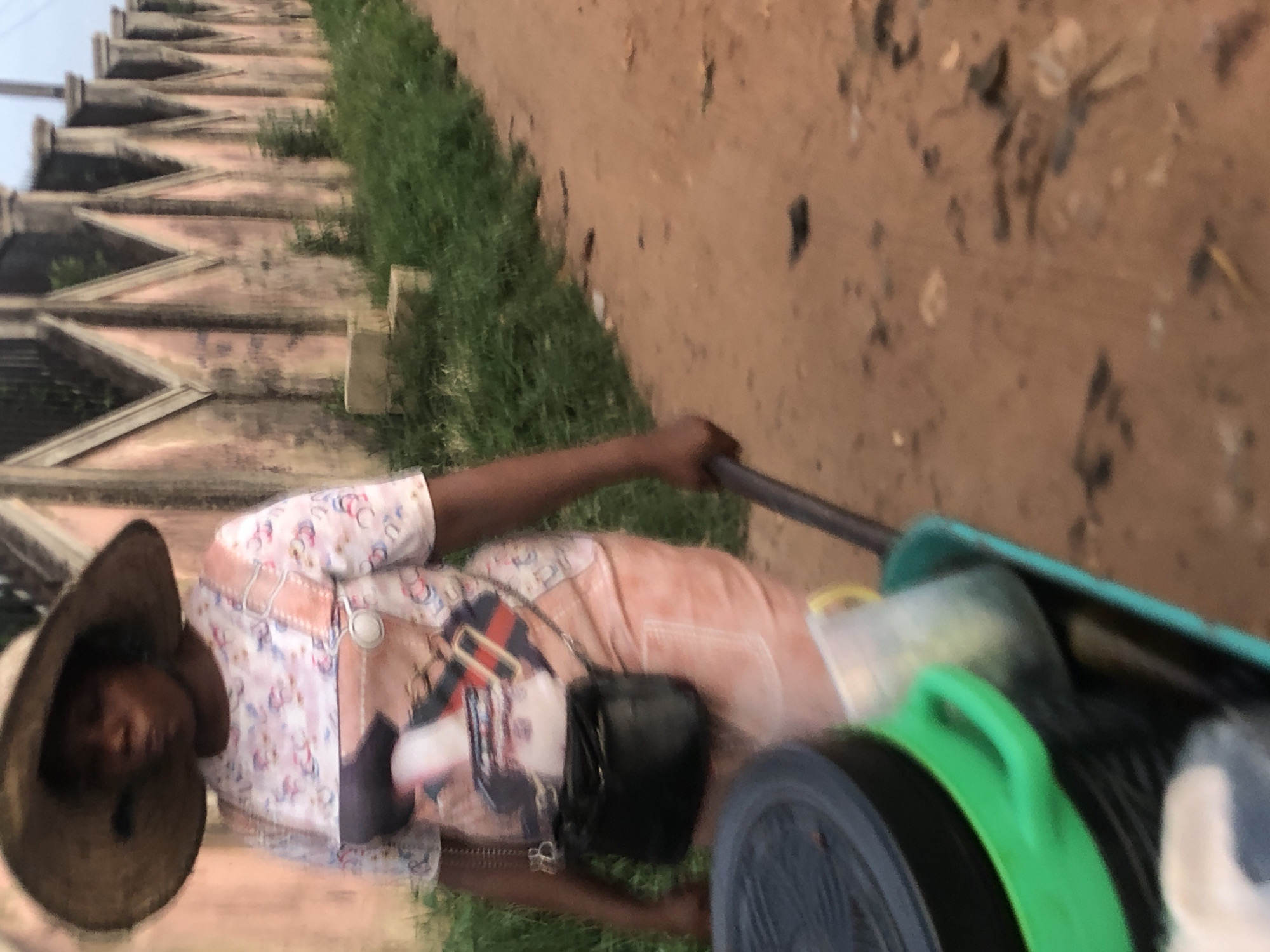 A person is pulling a cart on a dirt road next to a green strip of grass in what appears to be Nsugpe, Nigeria. The individual, wearing a straw hat and patterned shirt, has a black purse slung across their body and is holding onto the cart's handle. The cart itself is filled with various objects. To the left of the road and grass is a concrete wall with a sawtooth design. The ground is a mixture of brown dirt and debris.