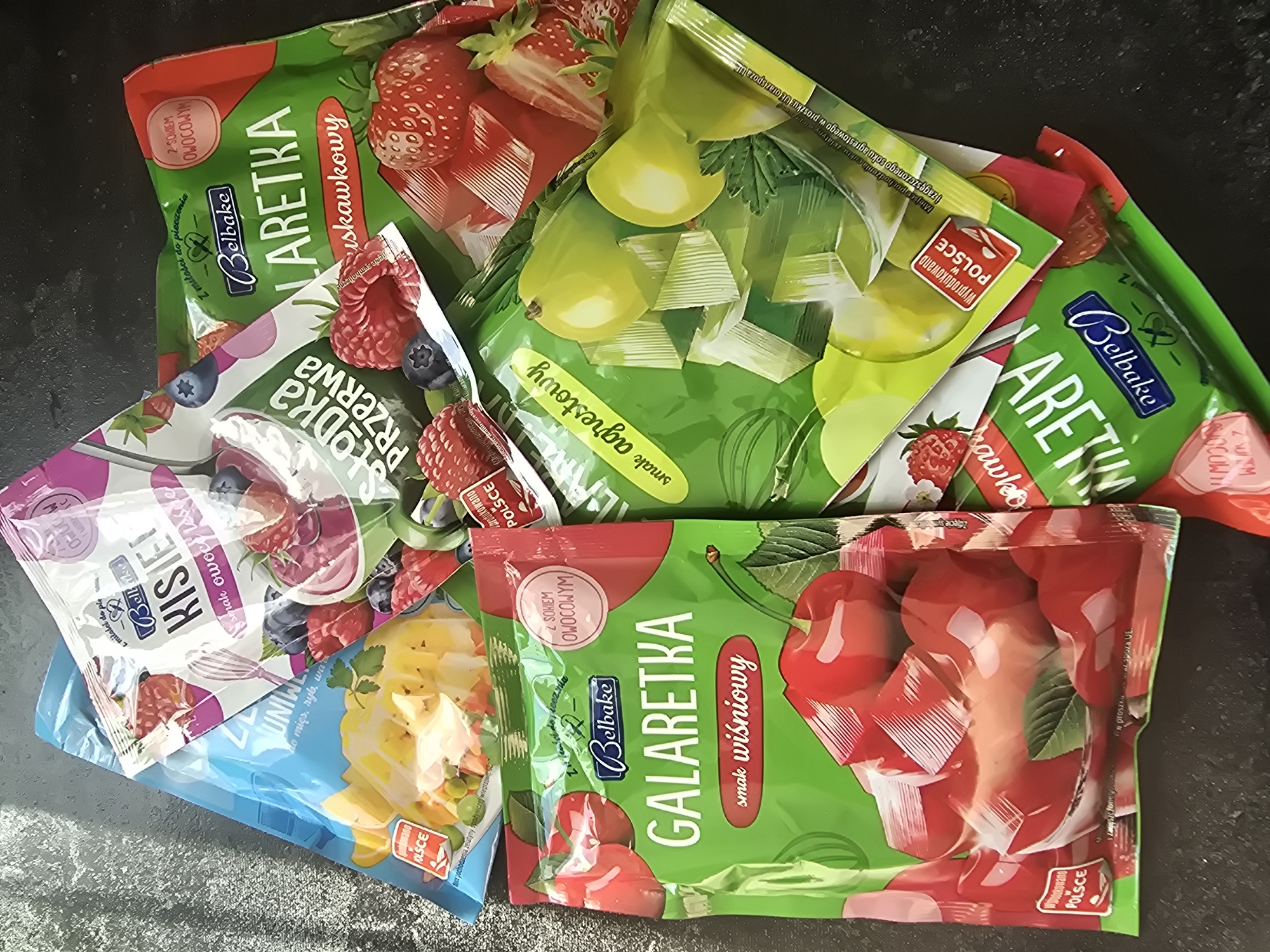 The image shows a pile of packages of "Galaretka," a type of jelly powder, on a dark, textured surface. The packages are mostly green, with some pink and blue accents, and feature colorful illustrations of fruits like strawberries, raspberries, blueberries, cherries, and what appear to be pears or green apples. The text on the packages is primarily in Polish, with brand names like "Belbake" and flavor descriptions such as "smak wiśniowy" (cherry flavor), "smak agrestowy" (gooseberry flavor), and "owoce leśne" (forest fruits). Some packages also indicate they are made in Poland ("Wyprodukowano w POLSCE"). The lighting suggests the photo was taken indoors, likely in daylight, as there are no strong shadows and the colors are well-illuminated. The arrangement of the packages is casual, with them overlapping and slightly scattered, implying they might be a collection or recently opened items. There are no people visible in the image.