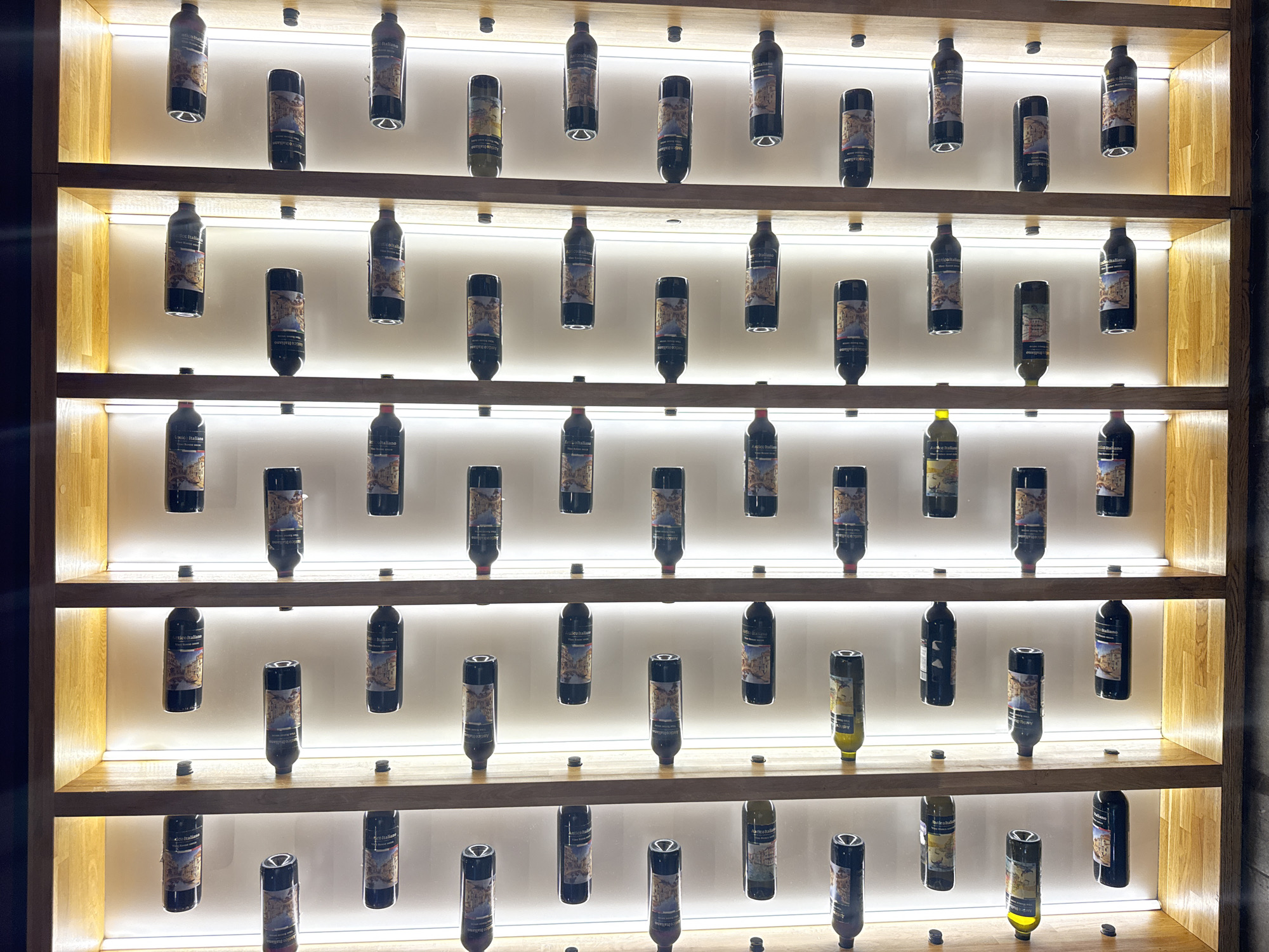 The image shows a vertical, multi-tiered wine rack with bottles of wine. The rack is made of light-colored wood and features individual compartments for each bottle, illuminated by interior lighting. The bottles are of various shapes and sizes, with labels visible on some. The wine rack appears to be in a well-lit indoor setting. This scene is located in Samara, Russia.