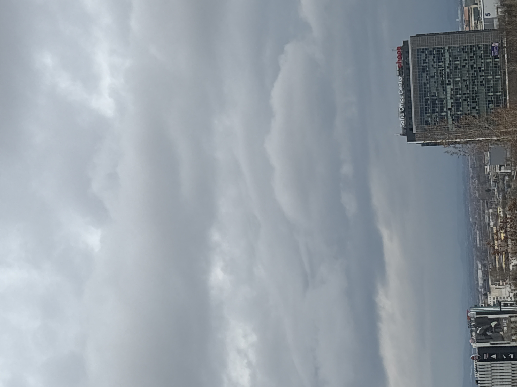 The image shows a cityscape of Sofia, Bulgaria, under a cloudy sky. Prominently visible is the Sofia Office Center building with the "shops" logo. Adjacent to it, other buildings are noticeable, including one with an advertisement featuring the words "TOO FAST". The overall environment is urban, and the atmospheric conditions appear overcast.