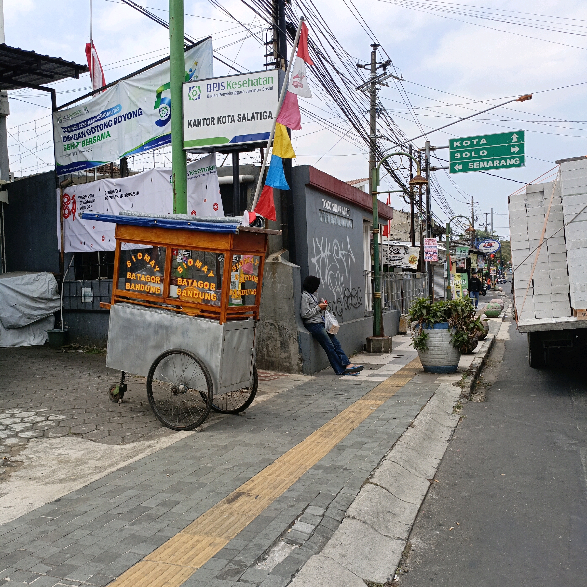 The image depicts a street scene in Salatiga, Indonesia, with a focus on a food cart and a person sitting by a wall. A mobile food cart, labeled "Siomay Batagor Bandung," is prominently positioned on the left side of the frame. It features a wooden top with glass display windows and a metal base. A person, dressed in a black head covering, grey long-sleeved shirt, and blue jeans, is seated against a grey wall on the right side of the cart, holding a phone and a plastic bag.
The environment includes a paved sidewalk with a yellow tactile paving strip, a street, and buildings. Various banners and signs are visible, including one for "BPJS Kesehatan" (Indonesian Health Social Security Agency) and another indicating "KANTOR KOTA SALATIGA" (Salatiga City Office). A green directional sign points towards "KOTA SOLO" and "SEMARANG." Overhead, a complex network of electrical wires and utility poles crisscrosses the sky. A truck loaded with white building materials is parked on the right side of the street. The overall scene suggests a commercial or public area within the city.