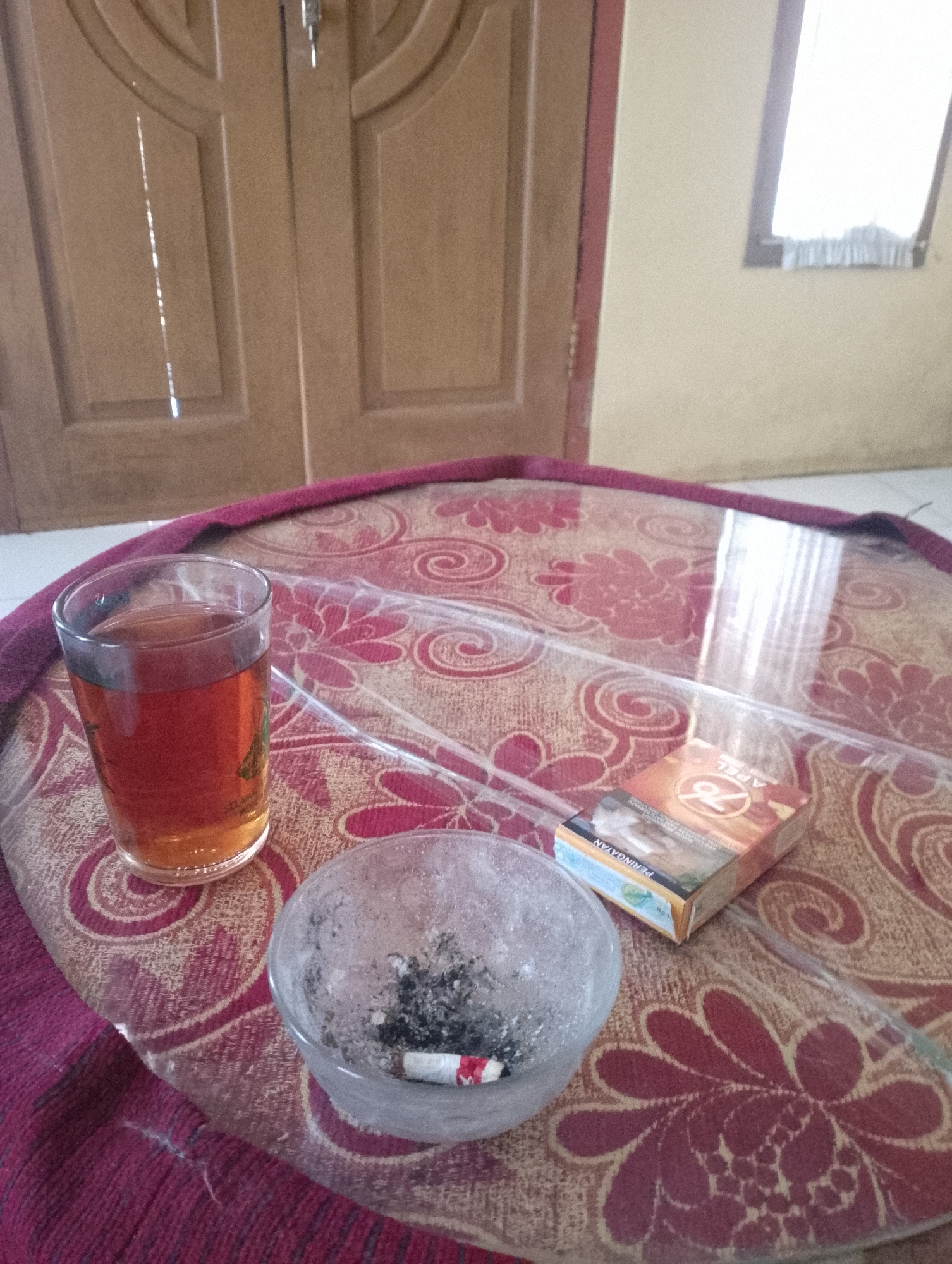 This image captures a still life scene on a circular table with a patterned cloth and a glass top. On the table, there is a glass of amber-colored liquid, likely tea, with a faint green design on its side. Adjacent to the glass is an ashtray containing cigarette ashes and a single cigarette butt. To the right of the ashtray and glass, a pack of cigarettes labeled "76 APEL" is positioned. The background shows a wooden door with vertical paneling and a small window with a light-colored curtain. The lighting suggests an indoor setting, possibly a home or a simple establishment. The overall impression is of a moment of pause, perhaps a break for a cigarette and tea. The presence of cigarettes and an ashtray suggests a smoking area, common in many Indonesian households and public spaces. Given the context of Sukamanah, Indonesia, this scene could be representative of a casual, everyday setting.